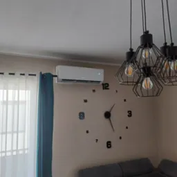 Biały klimatyzator na ścianie w salonie, obok okna z zasłonami i dużego zegara. W tle widoczna lampa i szara kanapa.