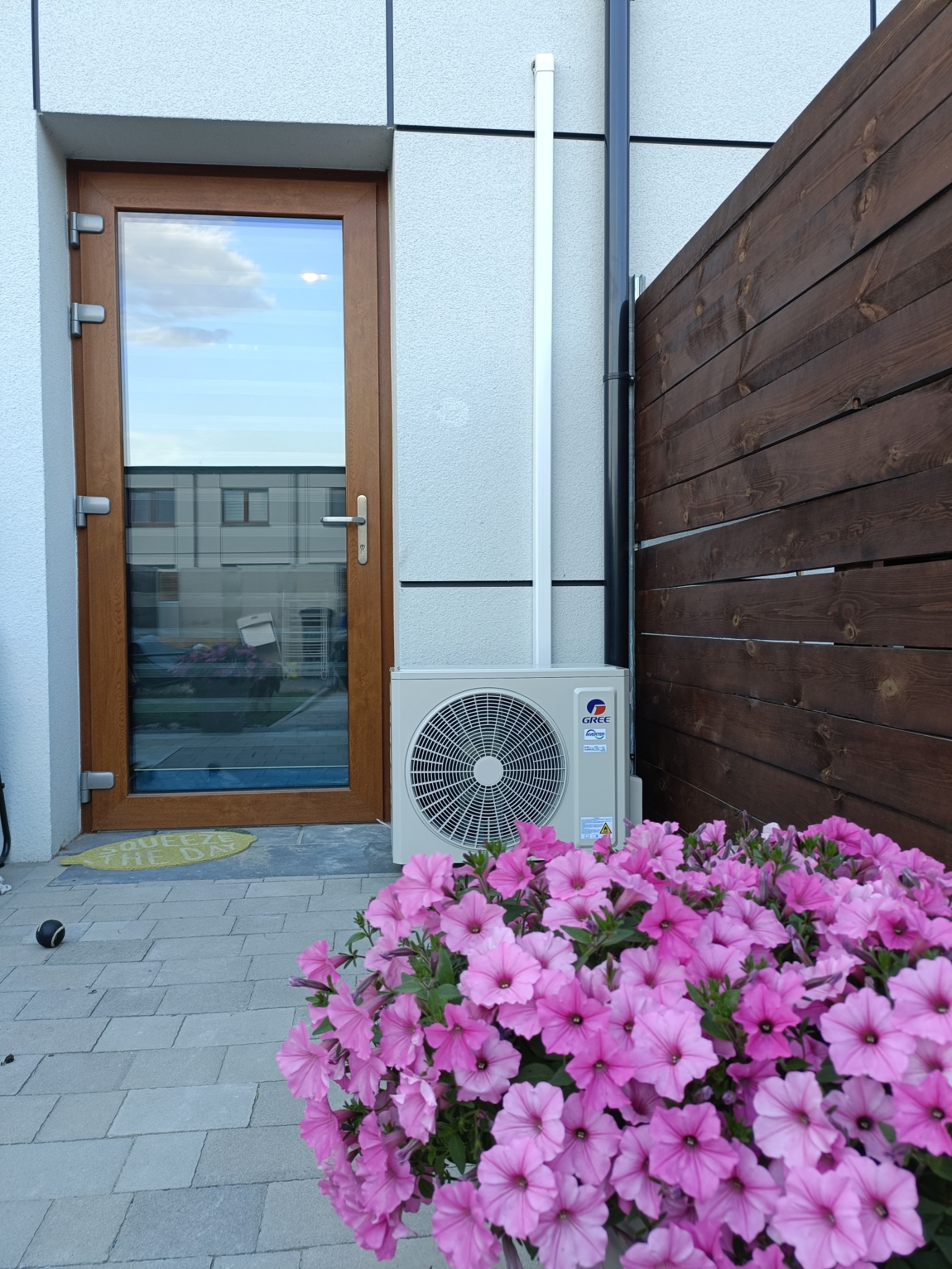 Biała jednostka klimatyzacji Gree obok brązowych drzwi wejściowych i drewnianego płotu, ozdobiona różowymi kwiatami na kamiennej kostce.