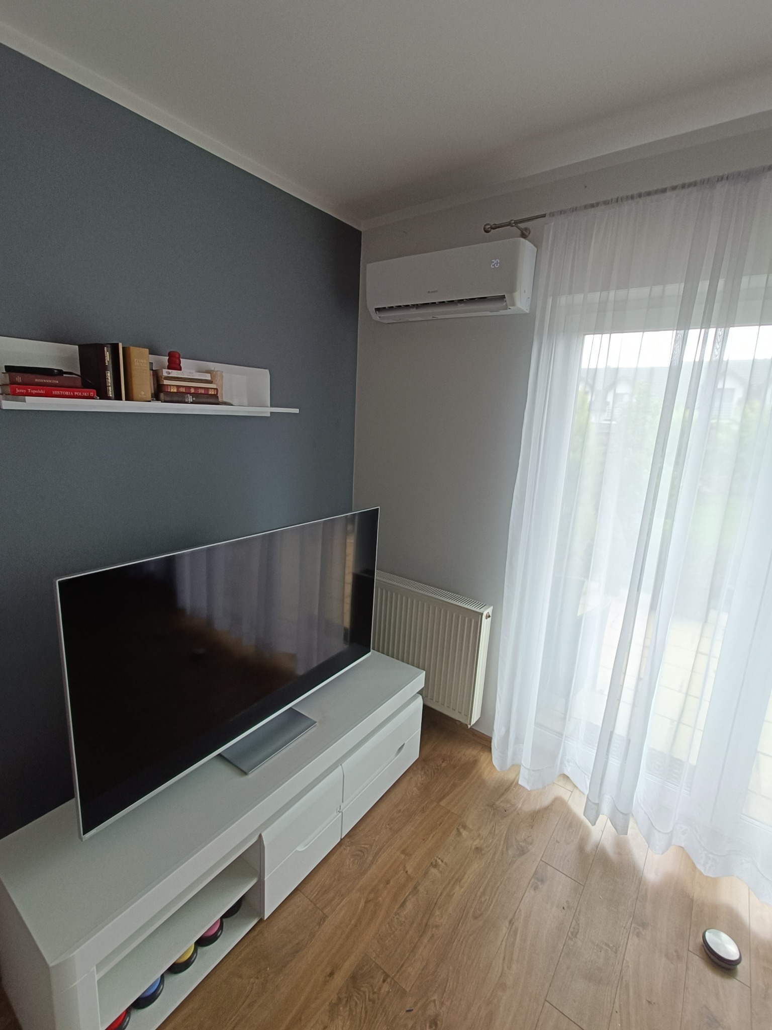 Nowoczesny salon z klimatyzatorem ściennym, białymi zasłonami i meblami. Widoczny telewizor, półka z książkami i grzejnik. Nowoczesne wnętrze z klimatyzacją.