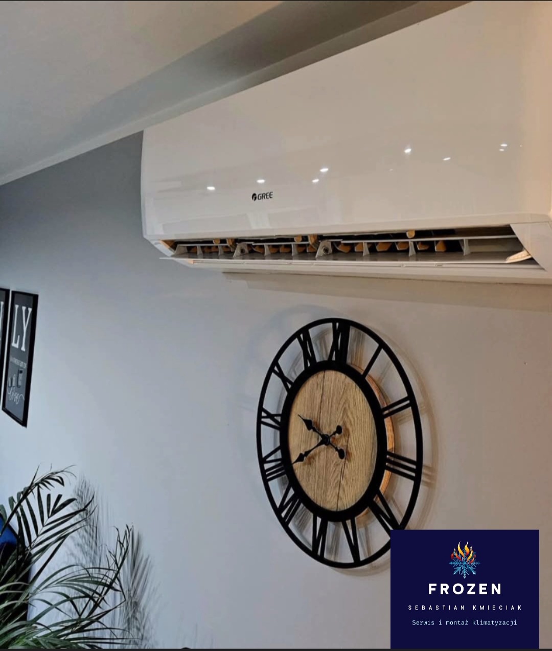 Biały klimatyzator marki Gree na ścianie w salonie, obok zegar w stylu retro i roślina doniczkowa. Widoczne logo firmy Frozen.