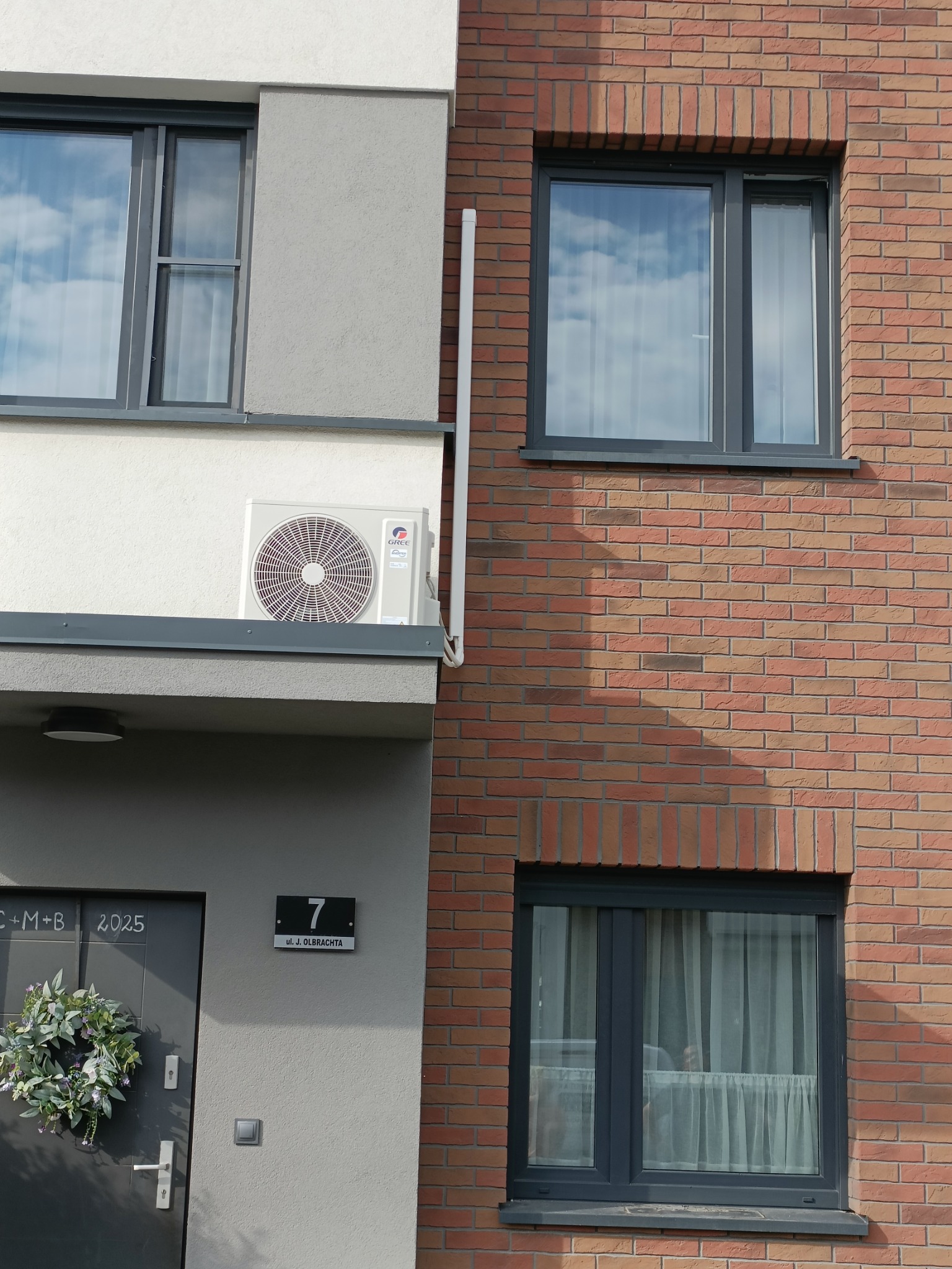 Biała jednostka zewnętrzna klimatyzacji Gree zamontowana na elewacji budynku obok okna i drzwi wejściowych z numerem 7 i dekoracyjnym wieńcem.