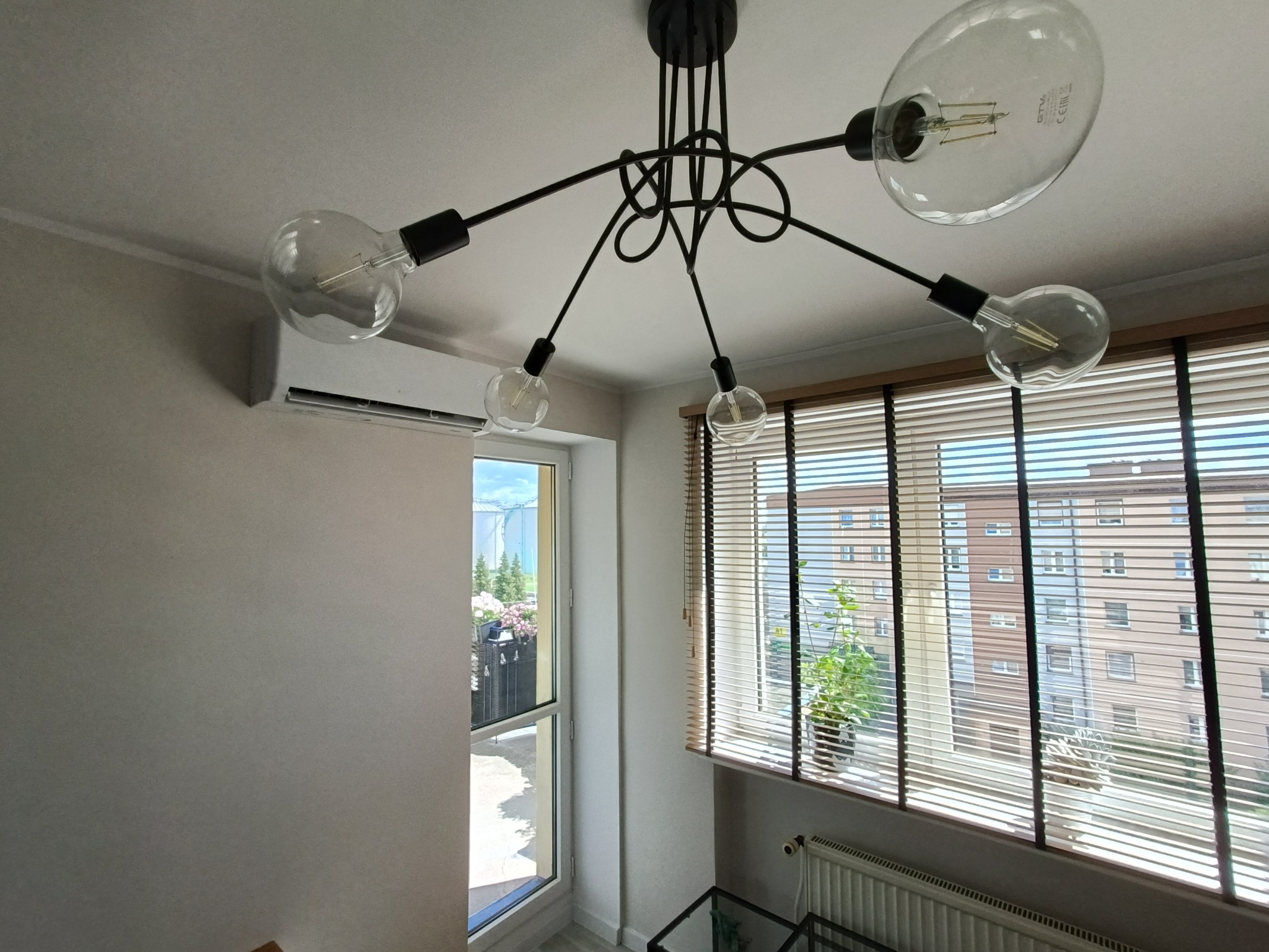 Zamontowany klimatyzator ścienny w jasnym wnętrzu, widoczny spod ozdobnej lampy sufitowej z żarówkami Edisona. W tle okno z żaluzjami.