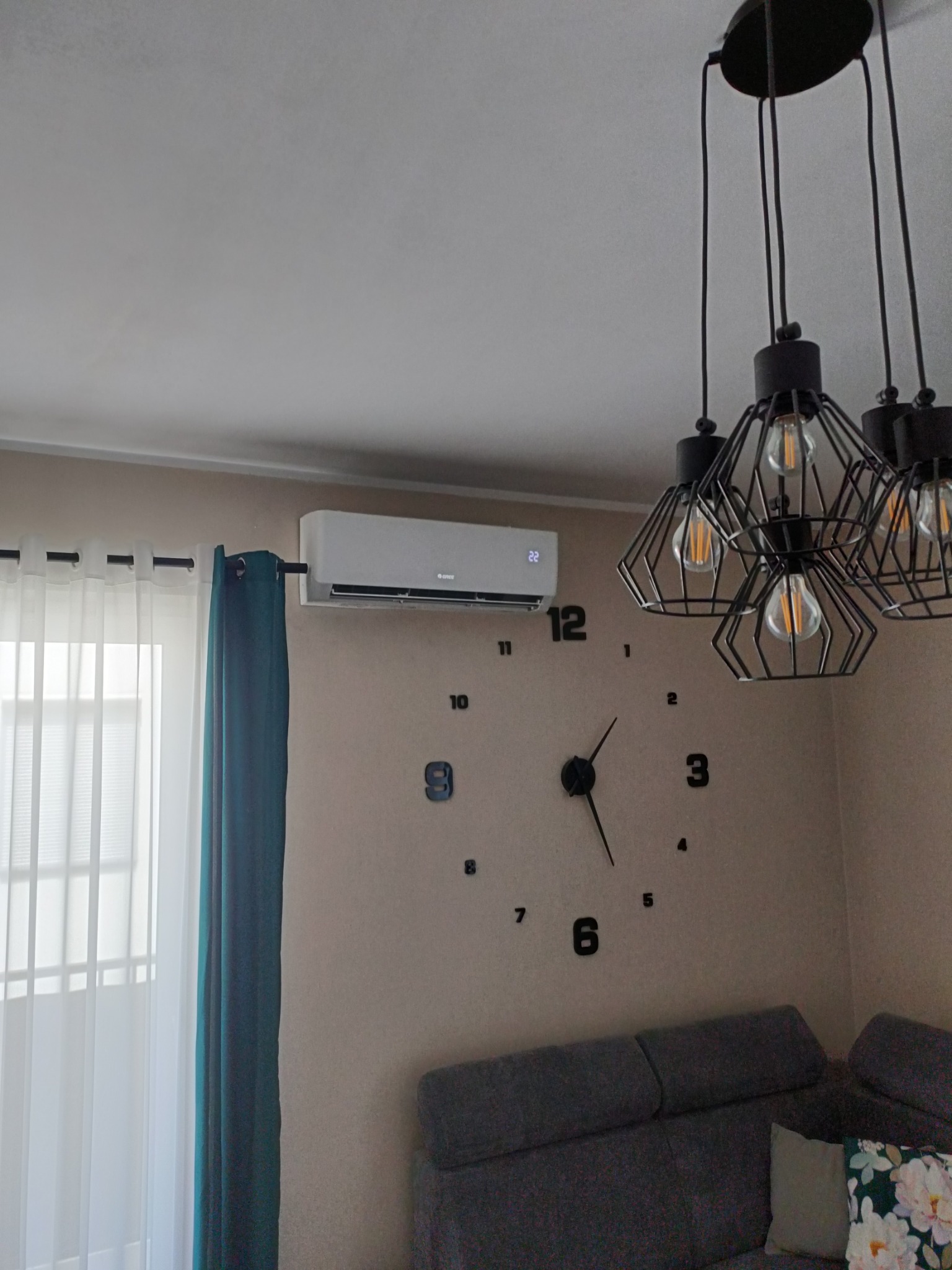 Biały klimatyzator na ścianie w salonie, obok okna z zasłonami i dużego zegara. W tle widoczna lampa i szara kanapa.