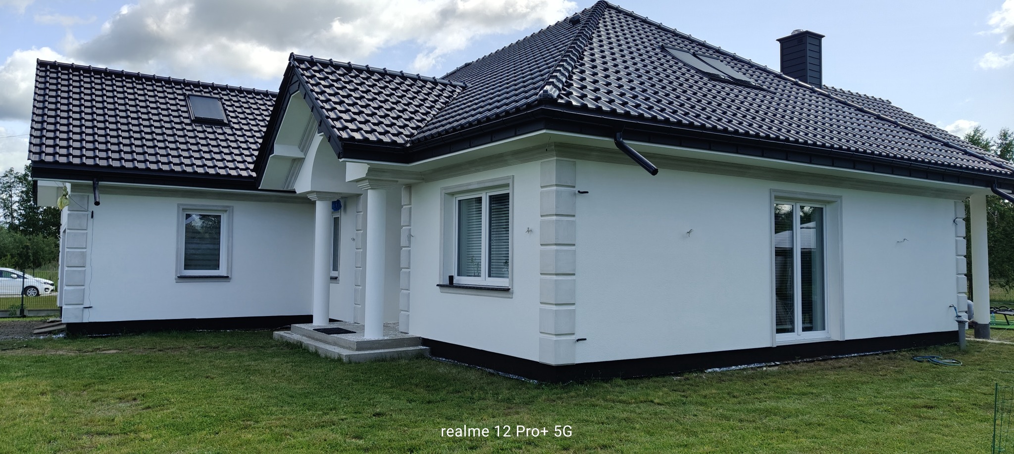 Biały dom jednorodzinny z ciemną dachówką, widok z zewnątrz. Elewacja z dekoracyjnymi elementami, kolumny przy wejściu, okna z roletami. Zadbany trawnik.
