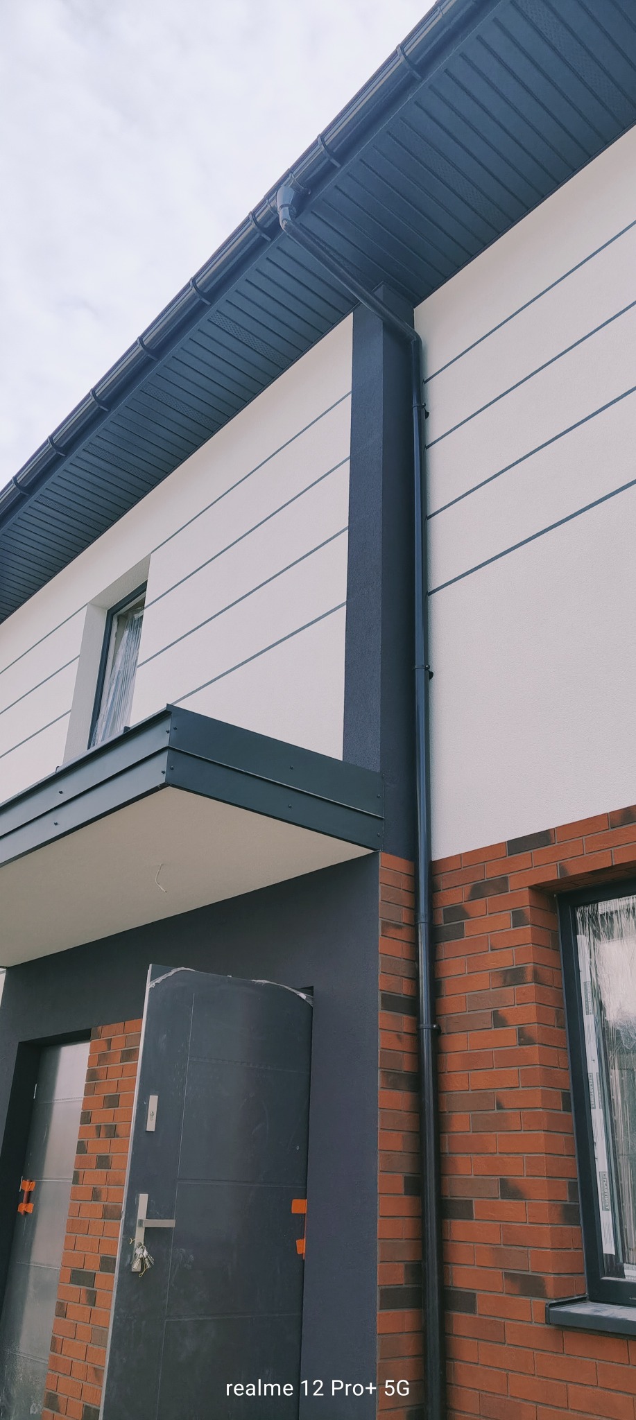 Nowoczesna elewacja domu z białym tynkiem, ciemnymi akcentami i ceglaną podstawą. Widoczny fragment dachu, okno i drzwi wejściowe. Ujęcie z dołu.