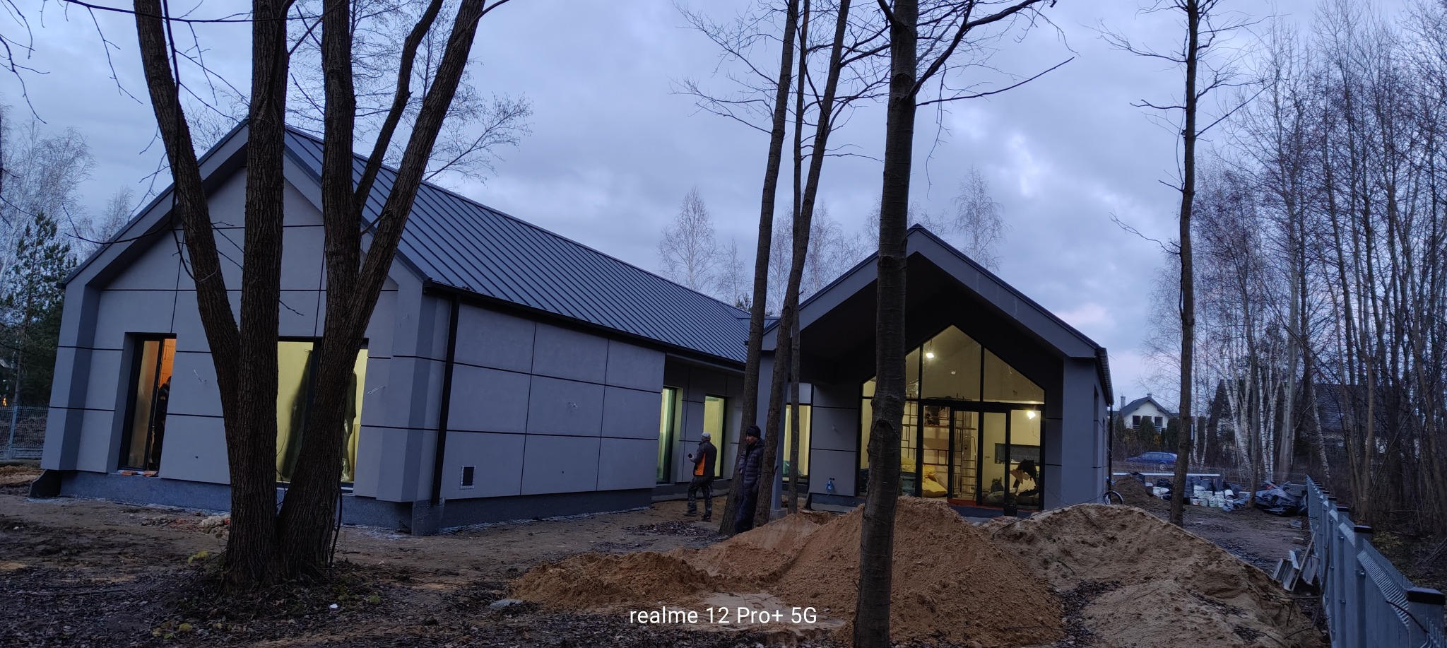 Nowoczesny dom z szarą elewacją panelową i ciemnym dachem, widoczni pracownicy, hałda piasku na pierwszym planie, drzewa bez liści w tle.
