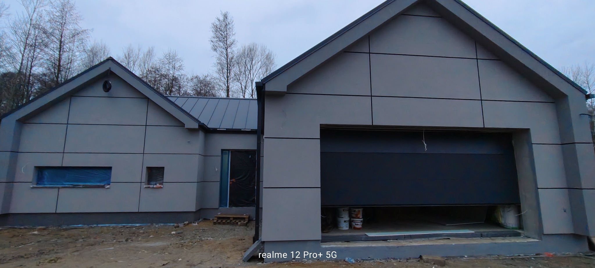 Nowoczesny dom z szarą elewacją panelową i garażem z częściowo otwartą bramą. Okna zabezpieczone folią. Widoczne puszki z farbą w garażu.