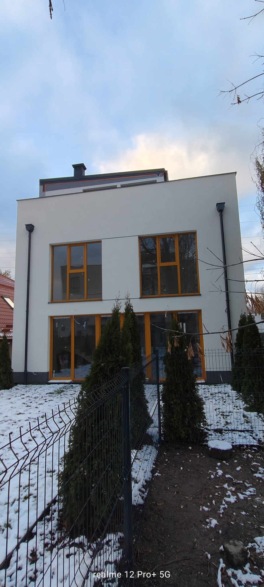 Dwupiętrowy dom z płaskim dachem i białymi ścianami, widoczny zza metalowego ogrodzenia w zimowej scenerii. Okna z drewnianymi ramami. Minimalistyczny styl.