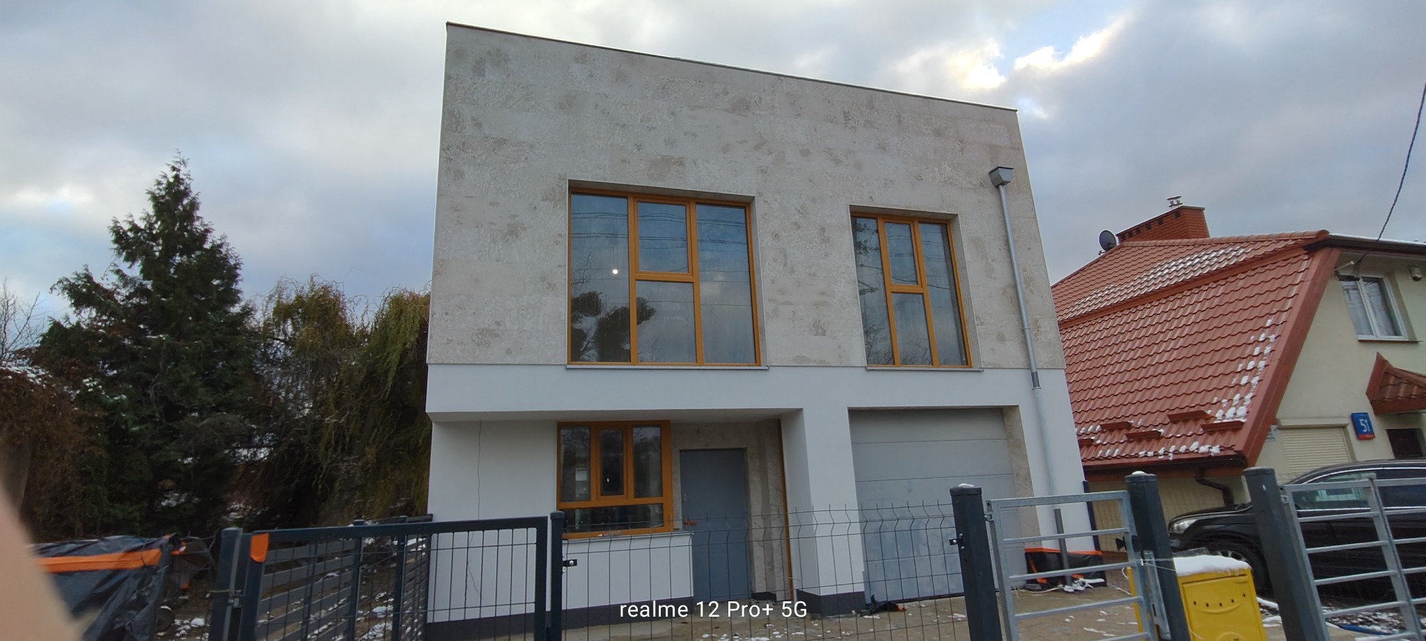 Dwukondygnacyjny dom z elewacją w odcieniach bieli i beżu, z drewnianymi ramami okiennymi, widziany zza metalowego ogrodzenia w pochmurny dzień.