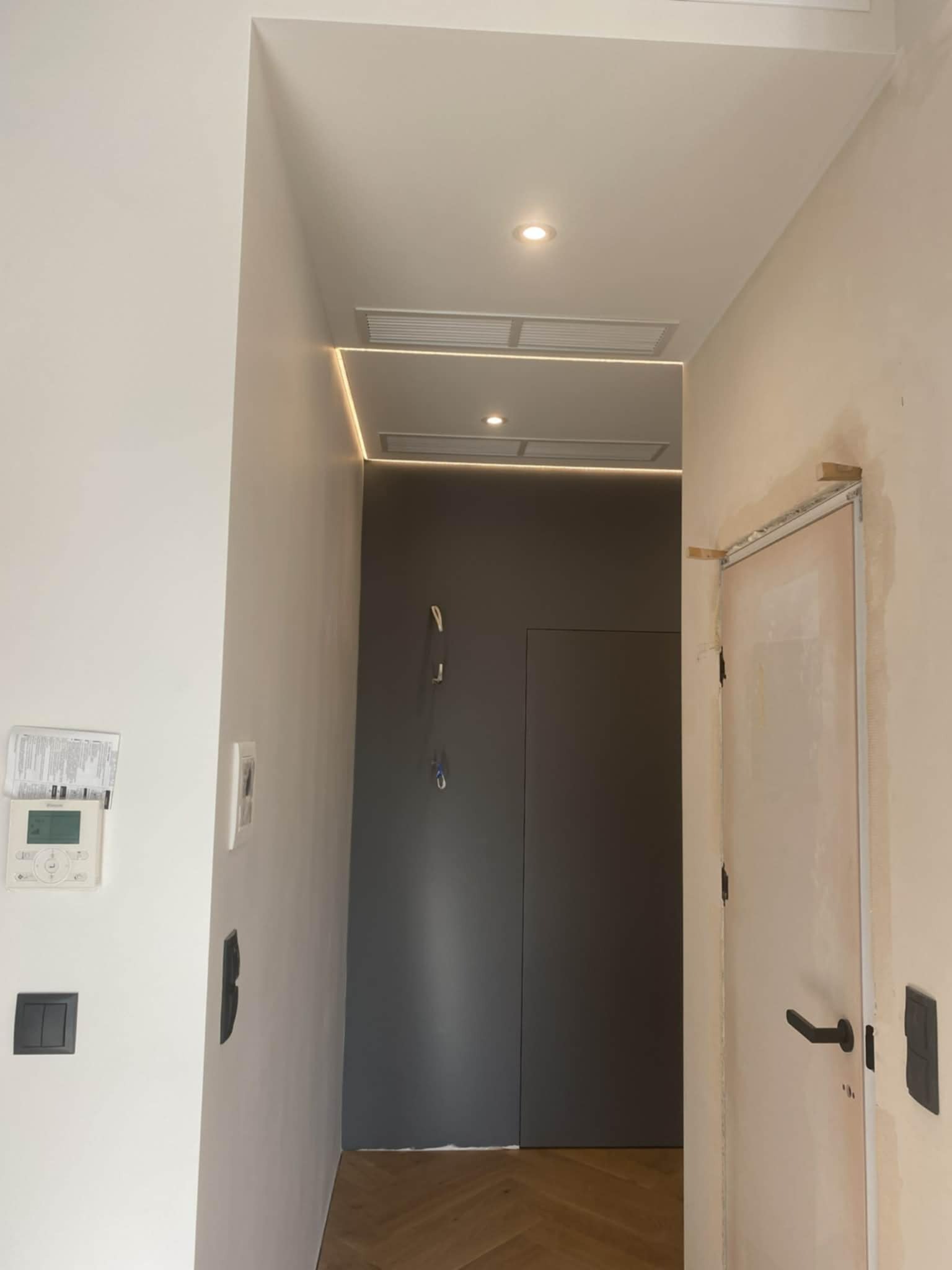 Wnętrze w trakcie remontu: widoczne przygotowane ściany, sufit z oświetleniem LED, zamontowane drzwi i elementy instalacji elektrycznej. Podłoga wyłożona parkietem.