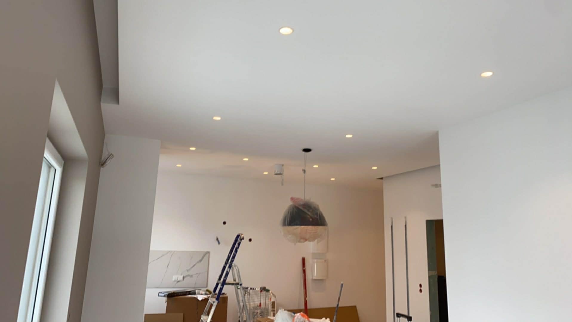Wnętrze po remoncie: białe ściany, sufit z oświetleniem punktowym, drabina i zabezpieczona lampa wisząca. Widoczne przygotowania do dalszych prac wykończeniowych.