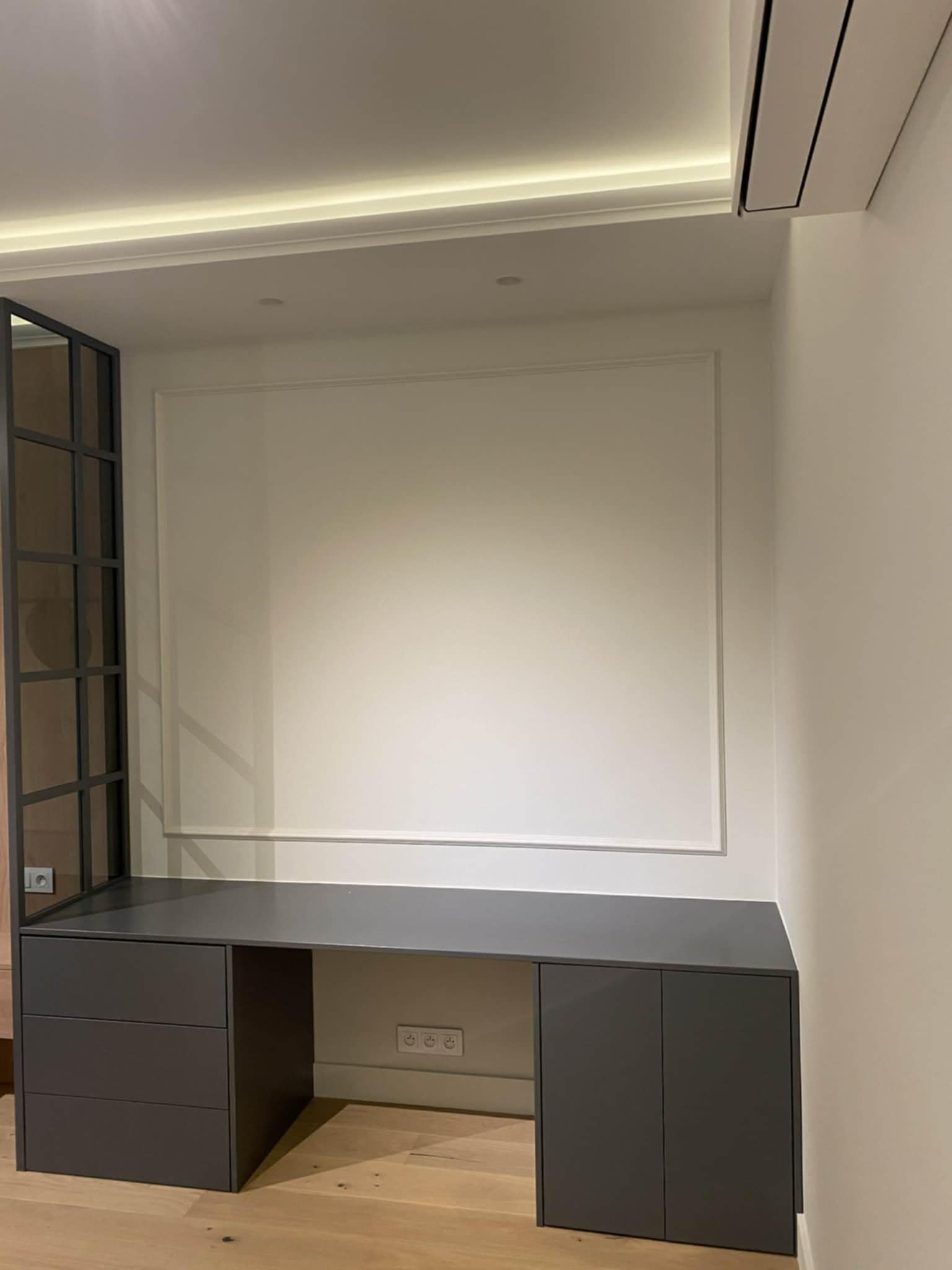 Szara zabudowa biurka z szufladami i szafkami, wkomponowana w białą ścianę z dekoracyjną ramą. Widoczna drewniana podłoga i fragment przeszklonej ścianki działowej.