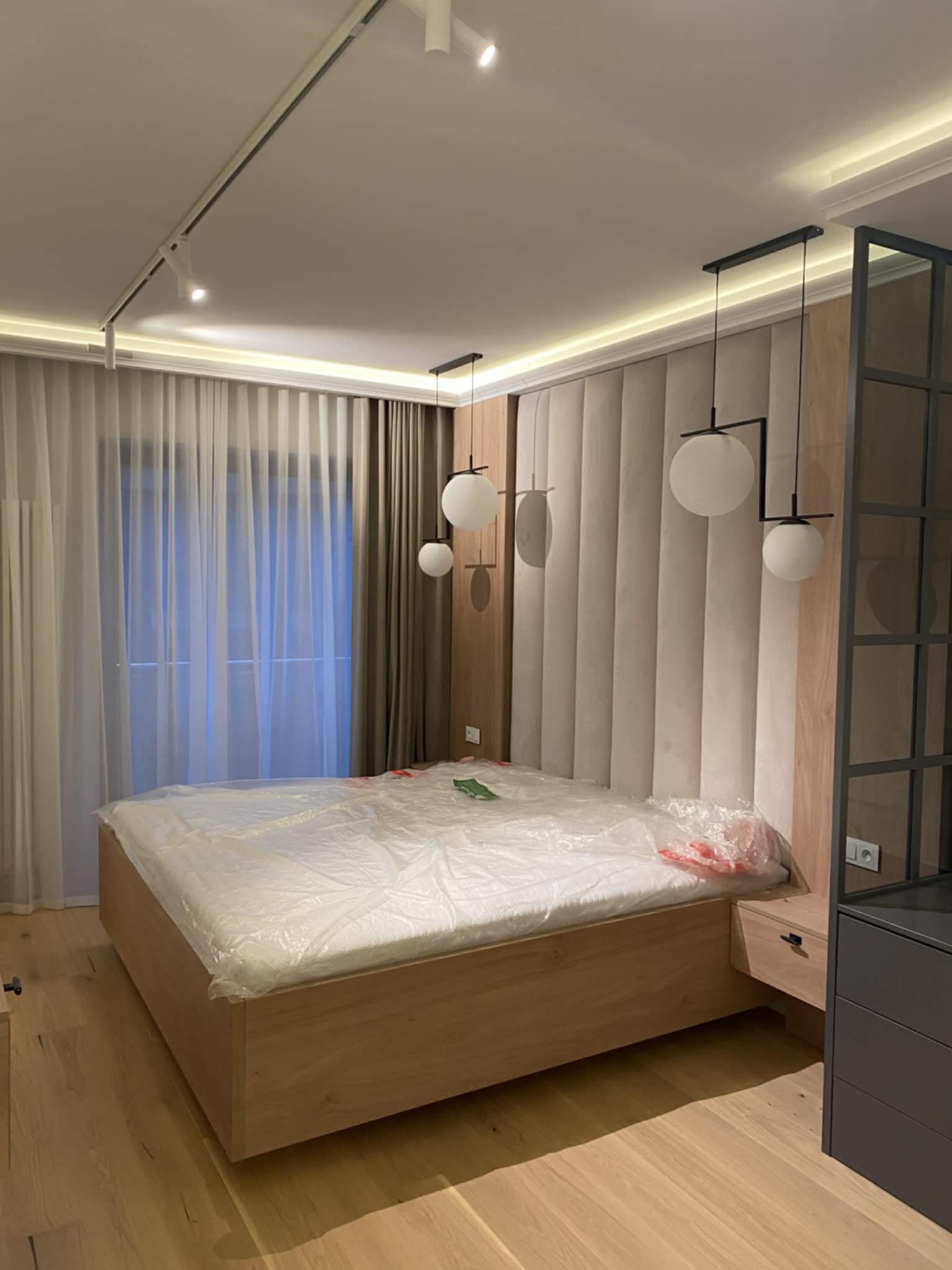 Nowoczesna sypialnia z łóżkiem w drewnianej ramie, tapicerowanym zagłówkiem i designerskimi lampami wiszącymi. Podłoga z jasnego drewna, okno z zasłonami.