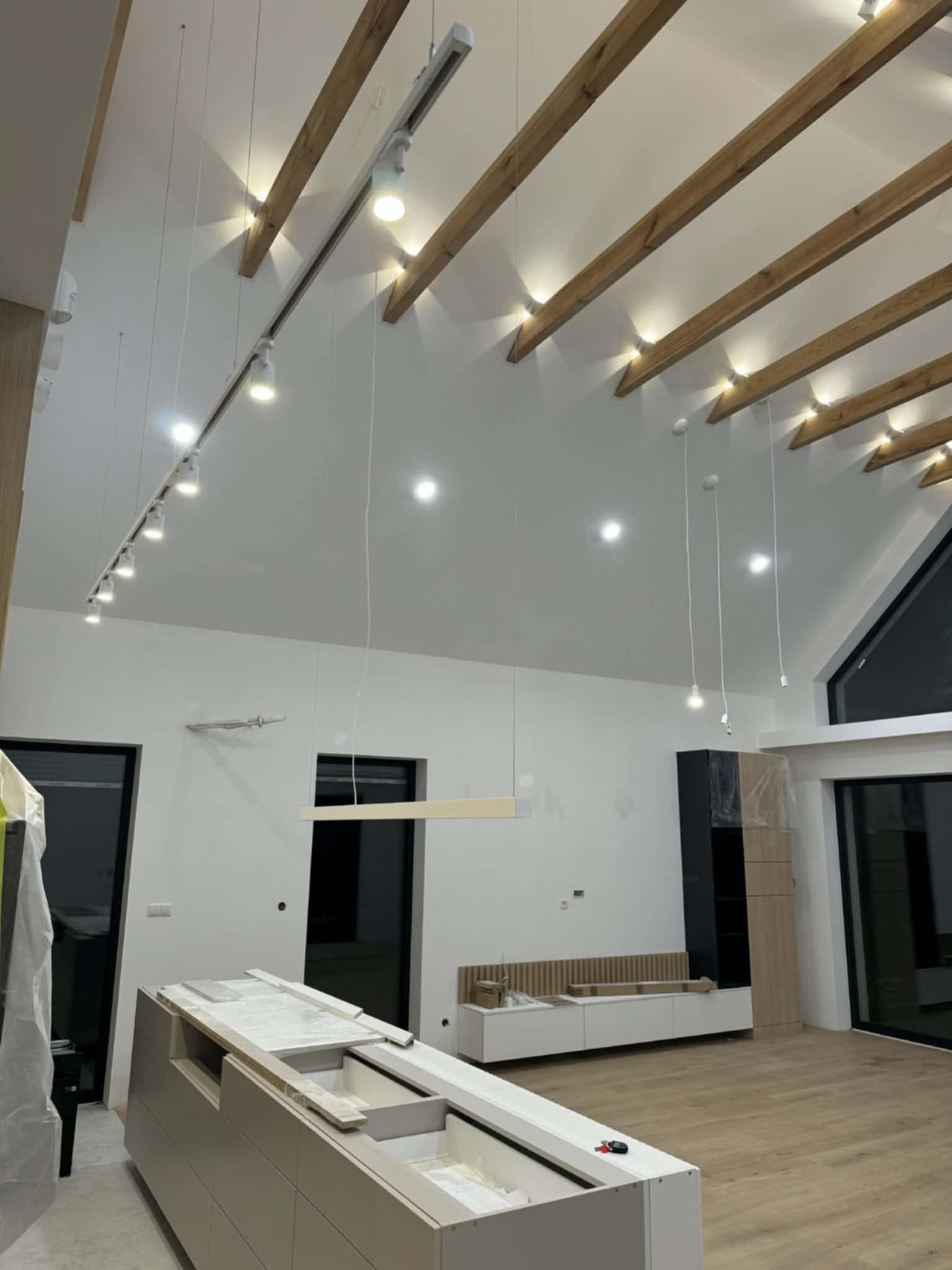 Wnętrze w trakcie wykańczania: drewniane belki stropowe z oświetleniem, białe ściany, okna, częściowo zmontowane meble kuchenne. Widoczne elementy instalacji elektrycznej.