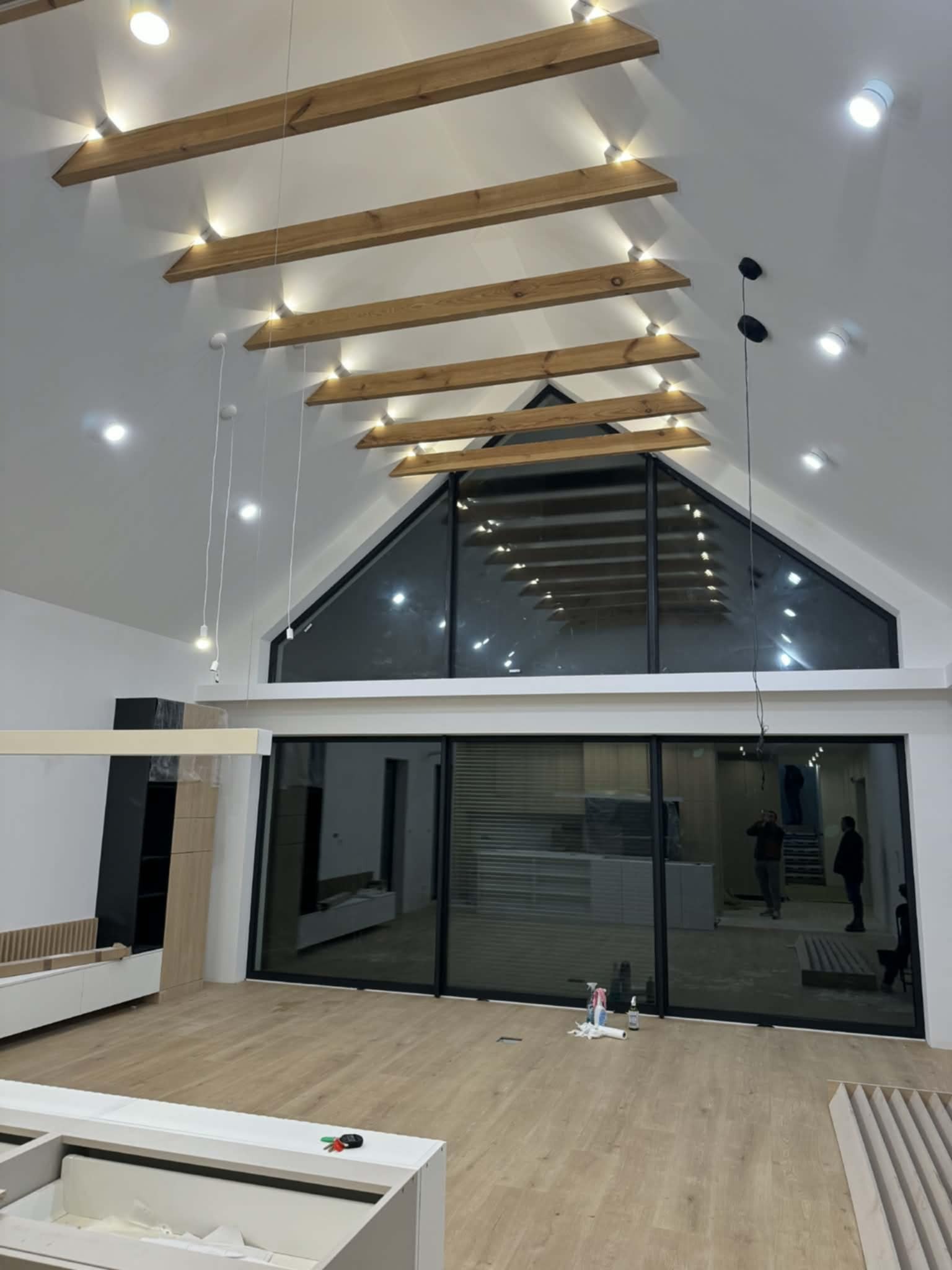 Wnętrze salonu z wysokim sufitem, drewnianymi belkami z oświetleniem, dużym oknem i przesuwnymi drzwiami. Widoczne elementy wykończenia i osoby w tle.