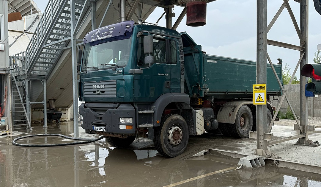 Ciężarówka MAN w punkcie mycia, z widocznym znakiem ostrzegawczym i elementami konstrukcji stalowej w tle. Widoczne ślady wody i błota na podłożu.