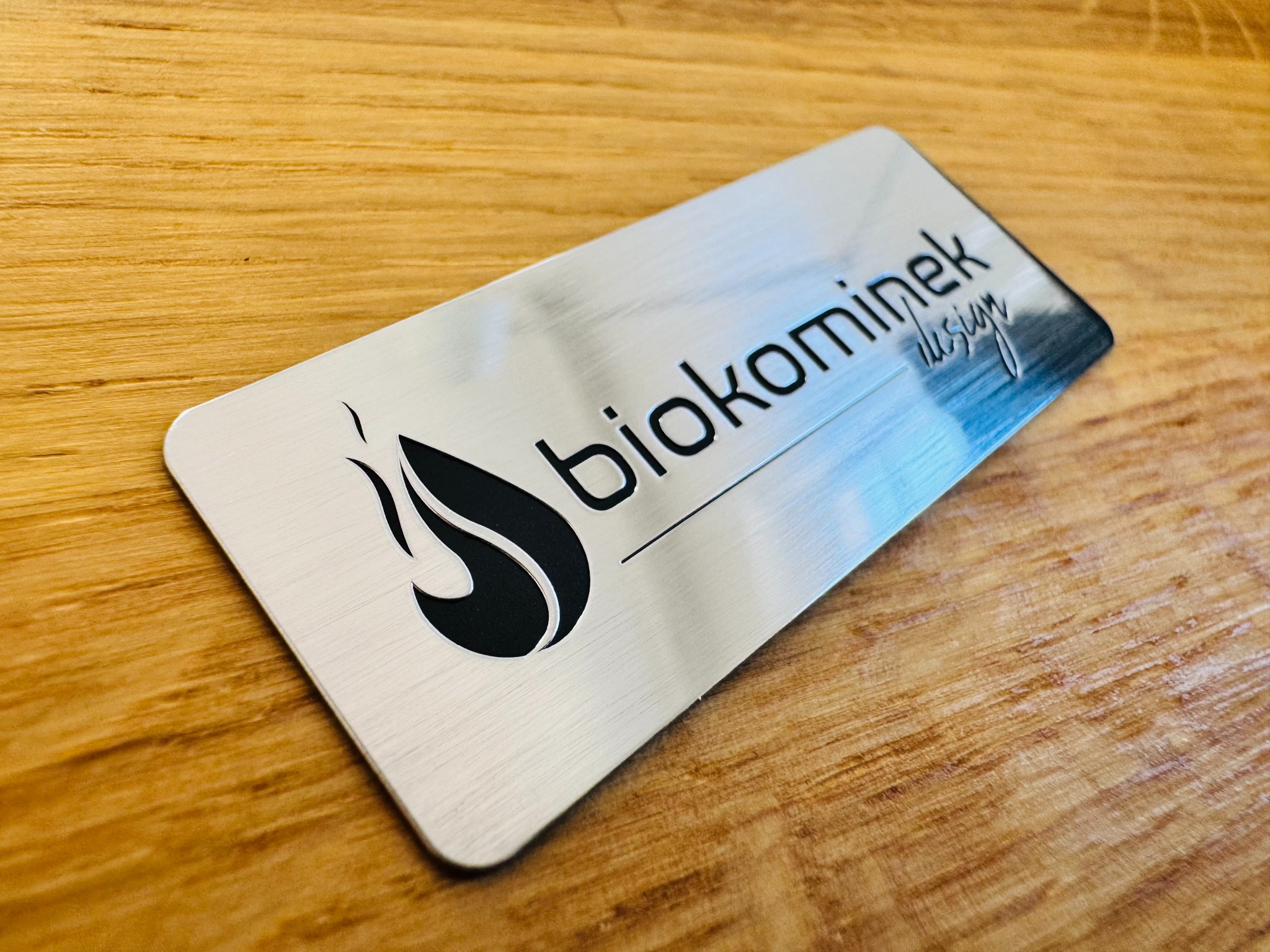 Metalowa, szczotkowana plakietka z logo 'biokominek design' leży na drewnianym blacie. Efekt chromu i mocny klej 3M nadają prestiż.