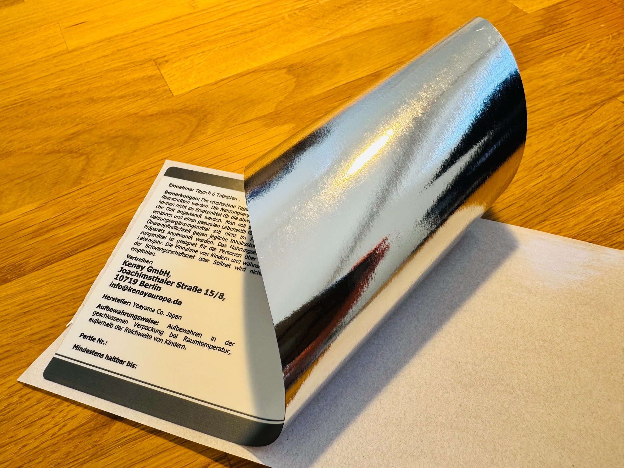 Srebrna etykieta samoprzylepna z nadrukiem na roli, leżąca na jasnym tle. Widoczny fragment tekstu w języku niemieckim na etykiecie.