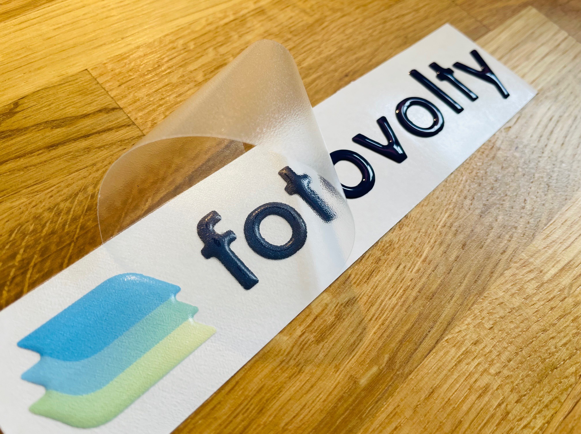 Elastyczna, wypukła naklejka z logo na białym tle, częściowo odklejona folia transportowa, widoczne trójwarstwowe logo i napis 'fotovolty' z efektem 3D.
