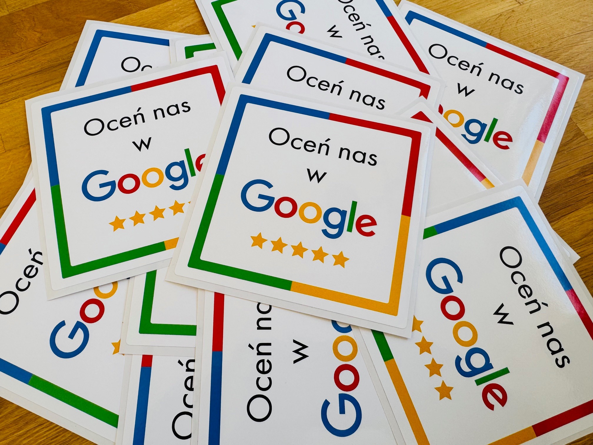 Stos kart z napisem 'Oceń nas w Google' i pięcioma gwiazdkami, z kolorowymi obwódkami (czerwona, zielona, żółta, niebieska), leżących na drewnianym blacie.