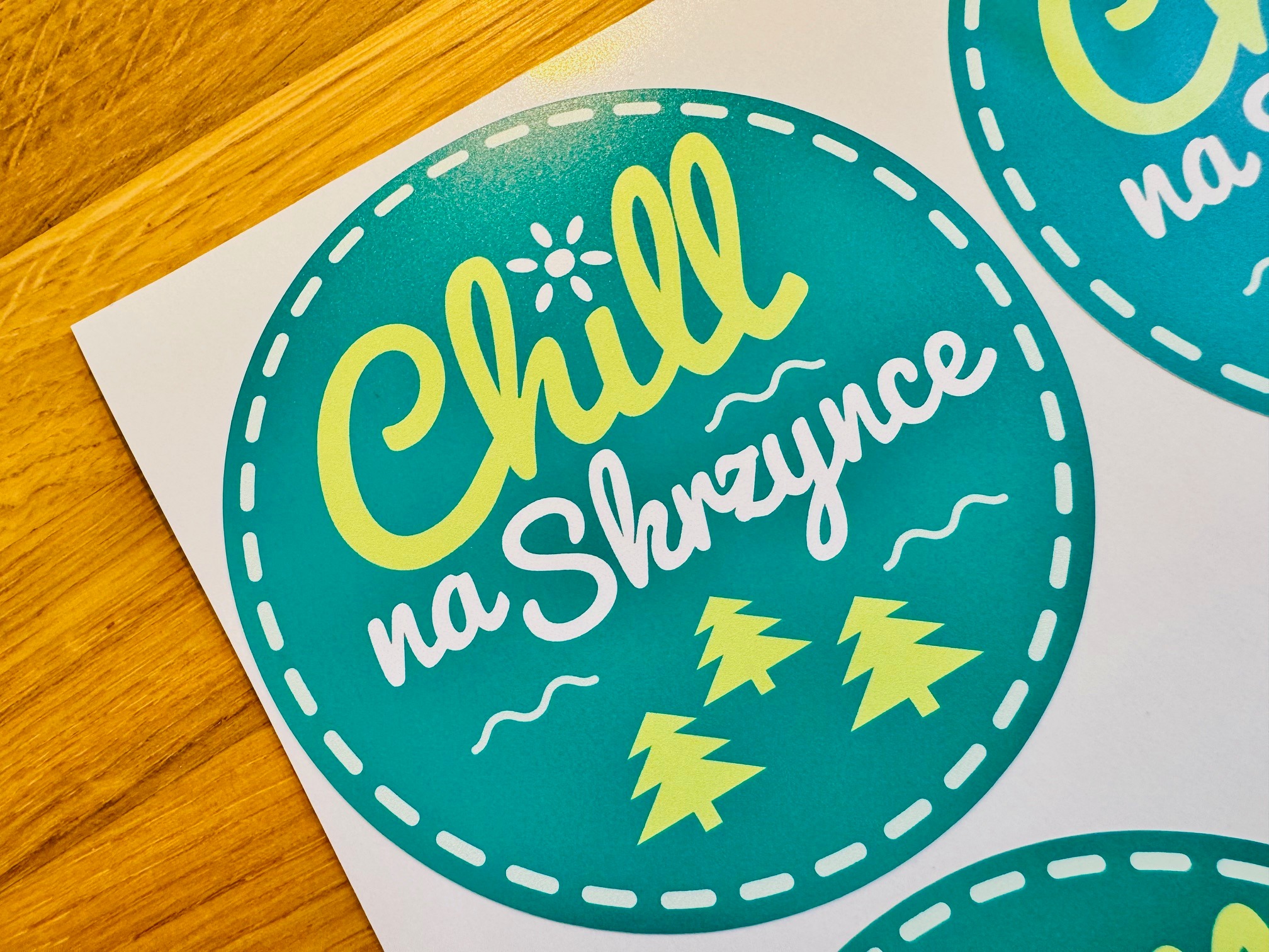 Druk na folii: okrągła, turkusowa etykieta z napisem 'Chill na Skrzynce' w kolorach żółtym i białym, ozdobiona motywem choinek. Widoczne krawędzie arkusza.
