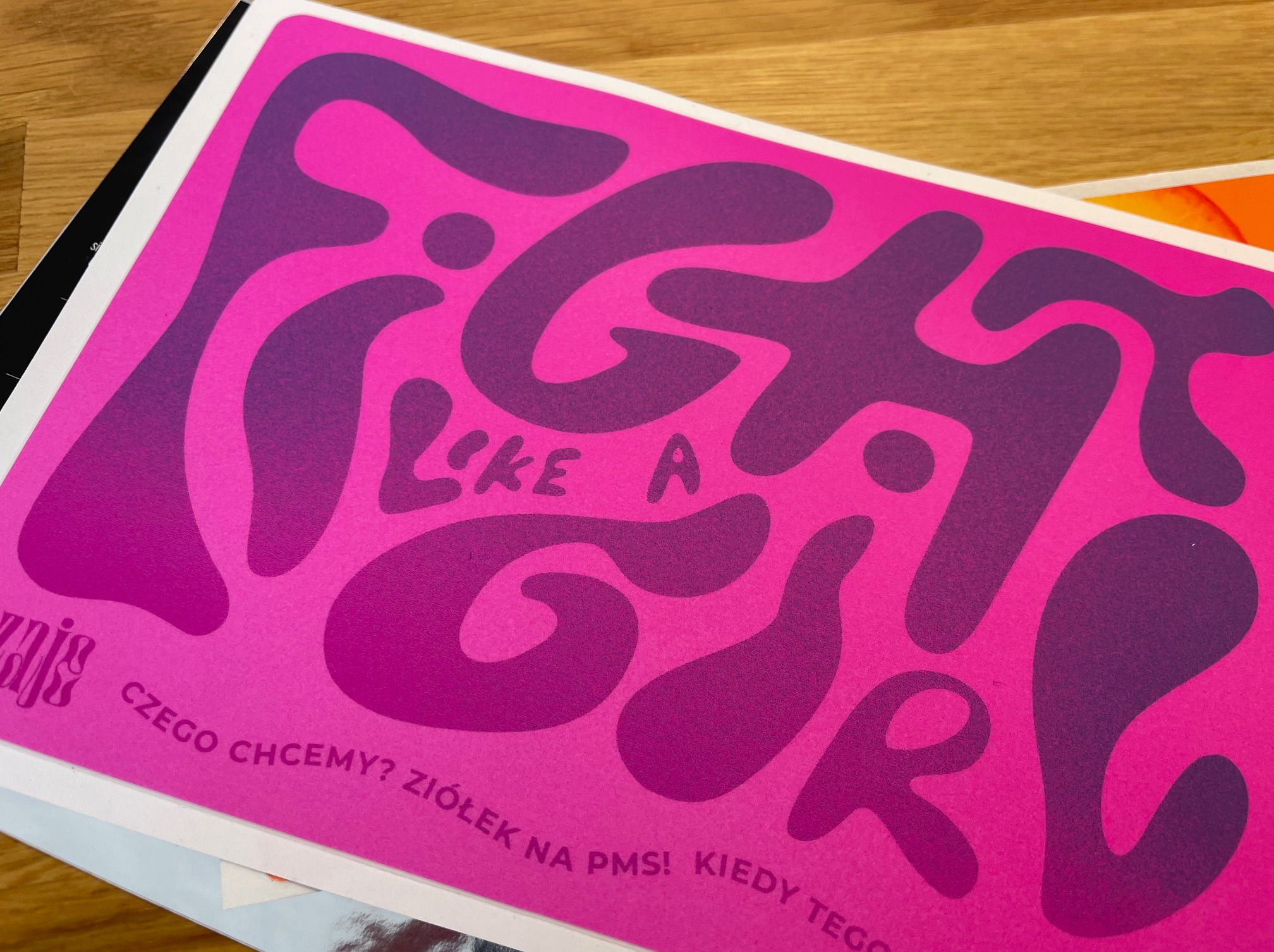 Różowo-fioletowa naklejka z hasłem 'Fight Like a Girl' w stylu retro, wykonana w Żninie. Matowe wykończenie, widoczne na drewnianym tle.