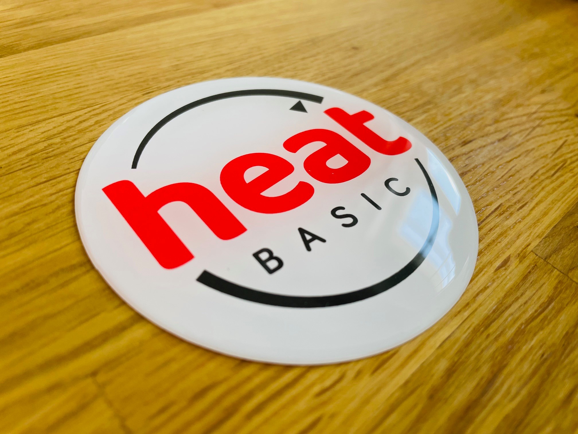 Biała, okrągła, wypukła etykieta z czerwonym napisem 'heat' i czarnym łukiem z napisem 'BASIC' na drewnianym blacie.