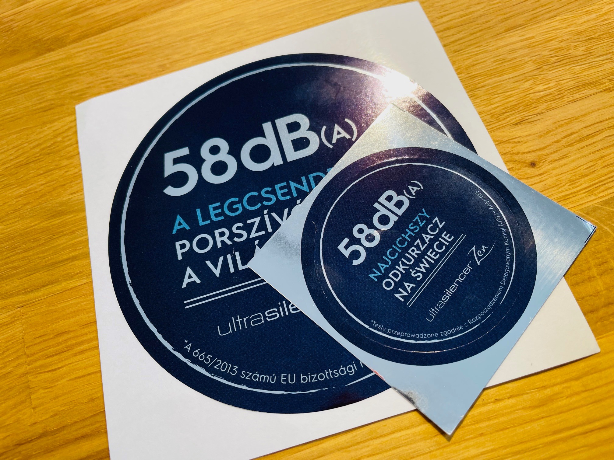 Dwie okrągłe, srebrne etykiety z ciemnoniebieskim nadrukiem, leżące na drewnianym blacie. Etykiety informują o poziomie hałasu 58dB(A) odkurzacza.