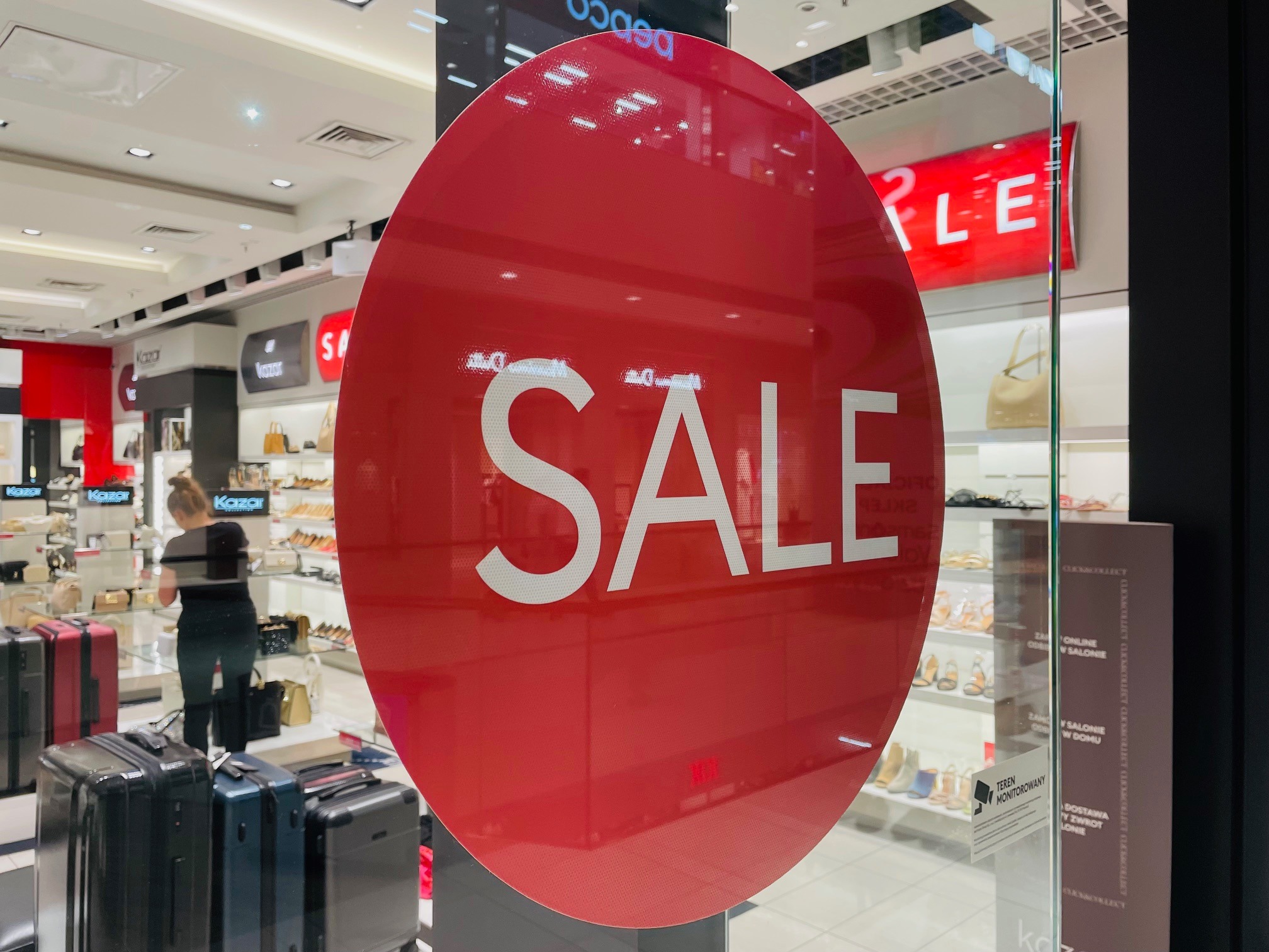 Naklejka wielkoformatowa SALE na szklanej witrynie salonu obuwniczego Kazor, z widocznymi walizkami i klientką w tle. Sezonowa reklama promocji.