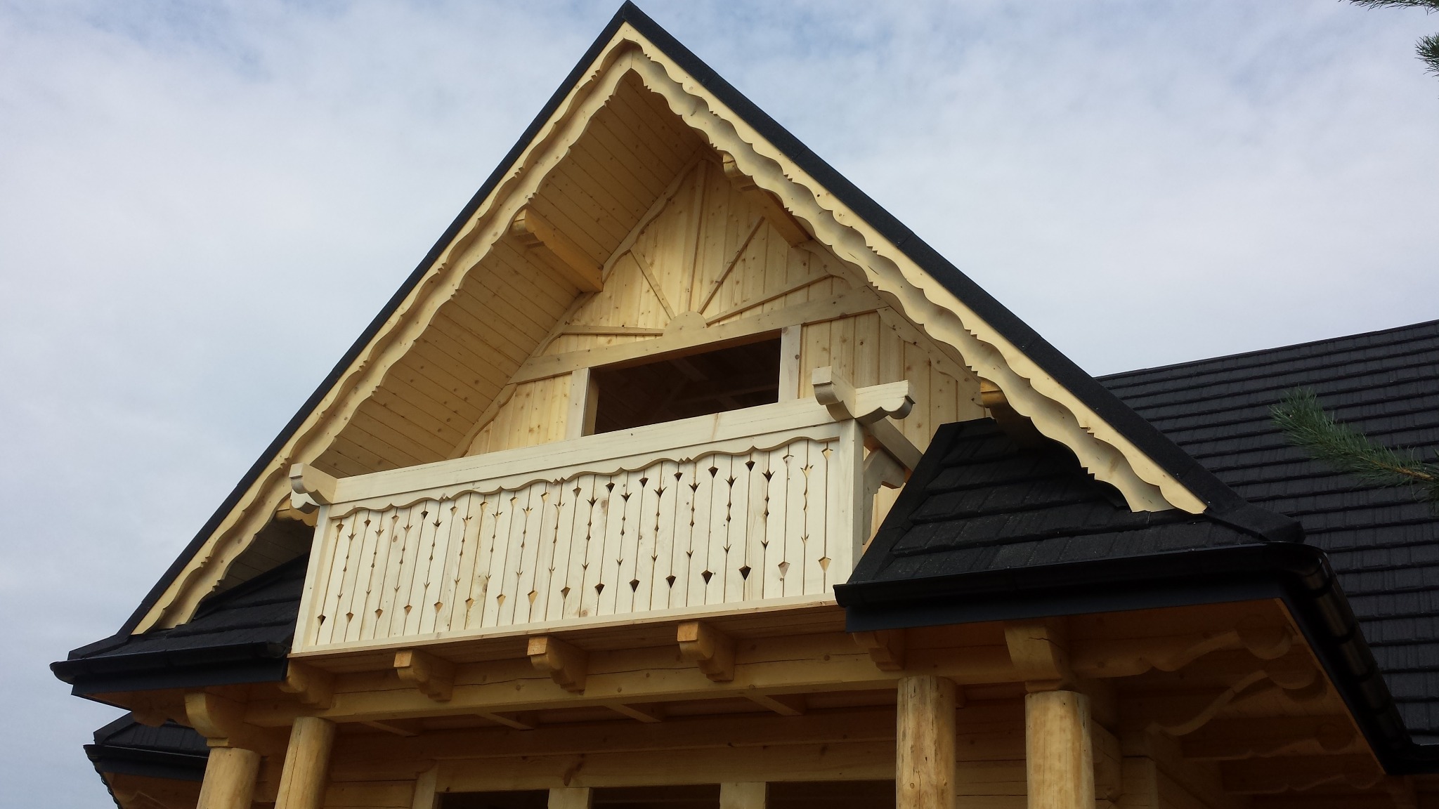 Fragment drewnianego domu z bali w stylu góralskim. Widoczny detal balkonu z ozdobną balustradą i wykończeniem dachu z ciemną dachówką. Surowe drewno i brak wykończenia.