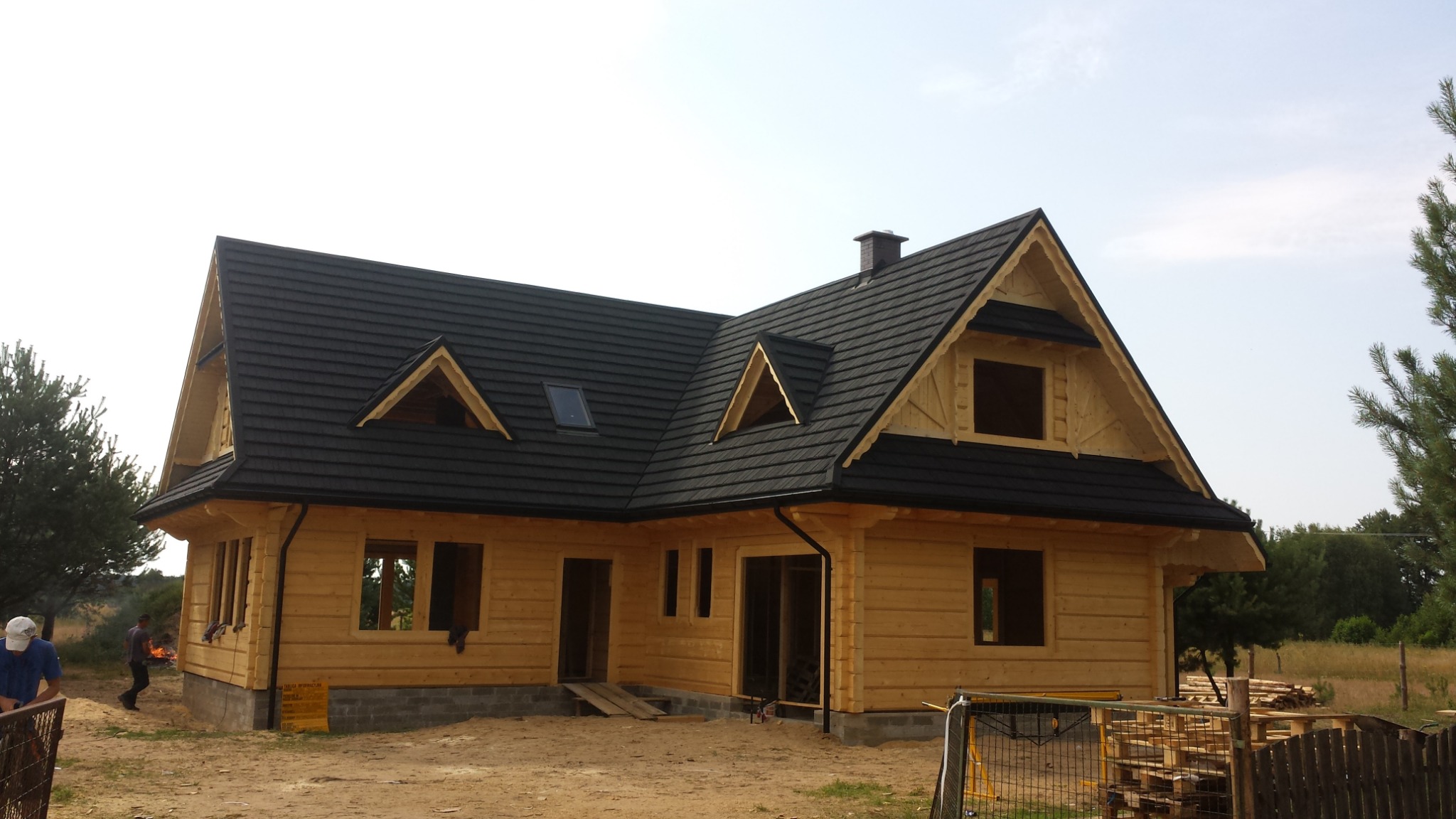 Surowy dom z bali z czarnym dachem, widoczne detale konstrukcyjne i brakujące okna. Na pierwszym planie prowizoryczne ogrodzenie i teren budowy.