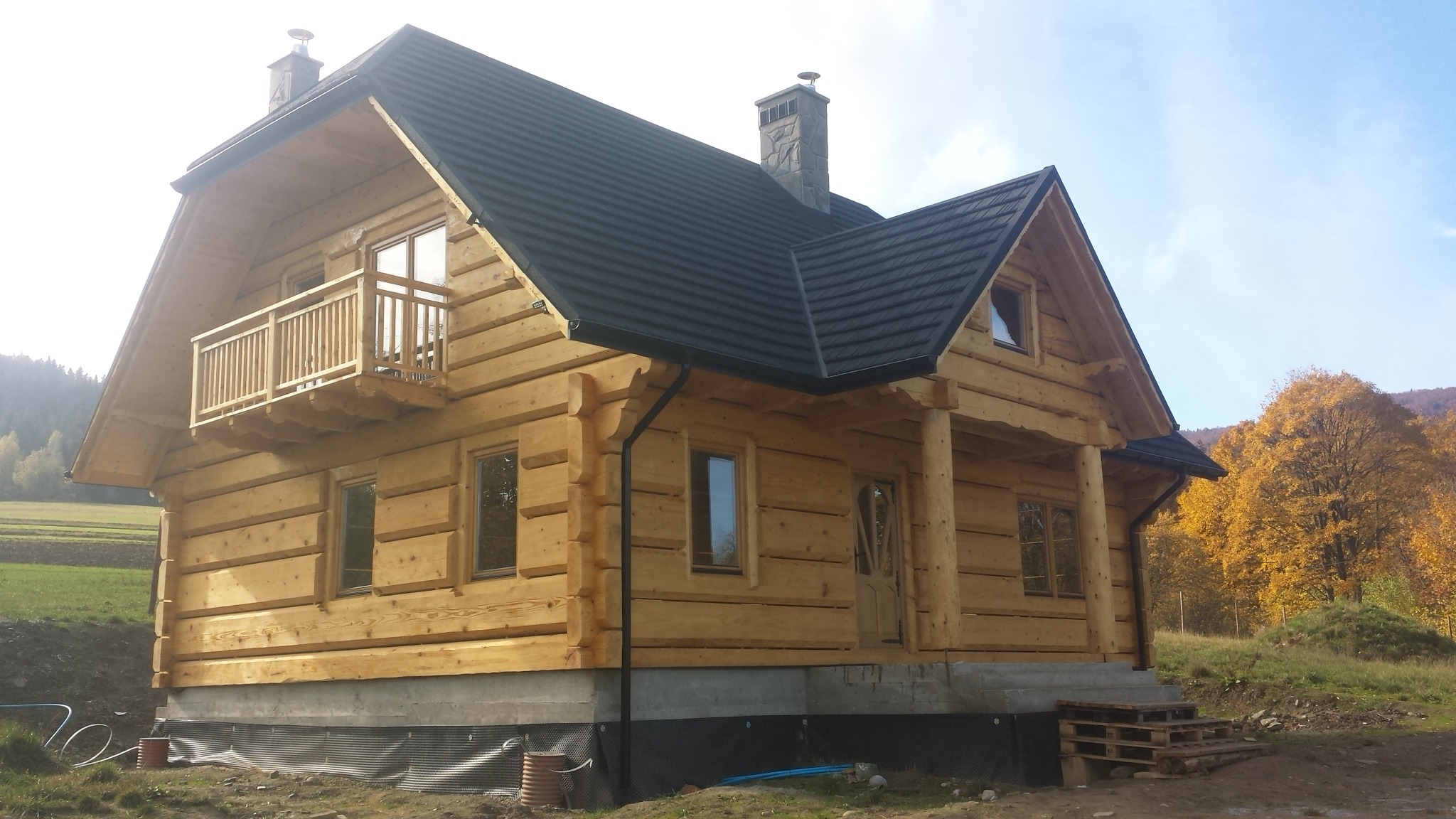Jasny dom z bali z balkonem i ciemnym dachem na tle jesiennego krajobrazu. Widoczne detale konstrukcyjne i naturalne materiały. Nierówny teren wokół.