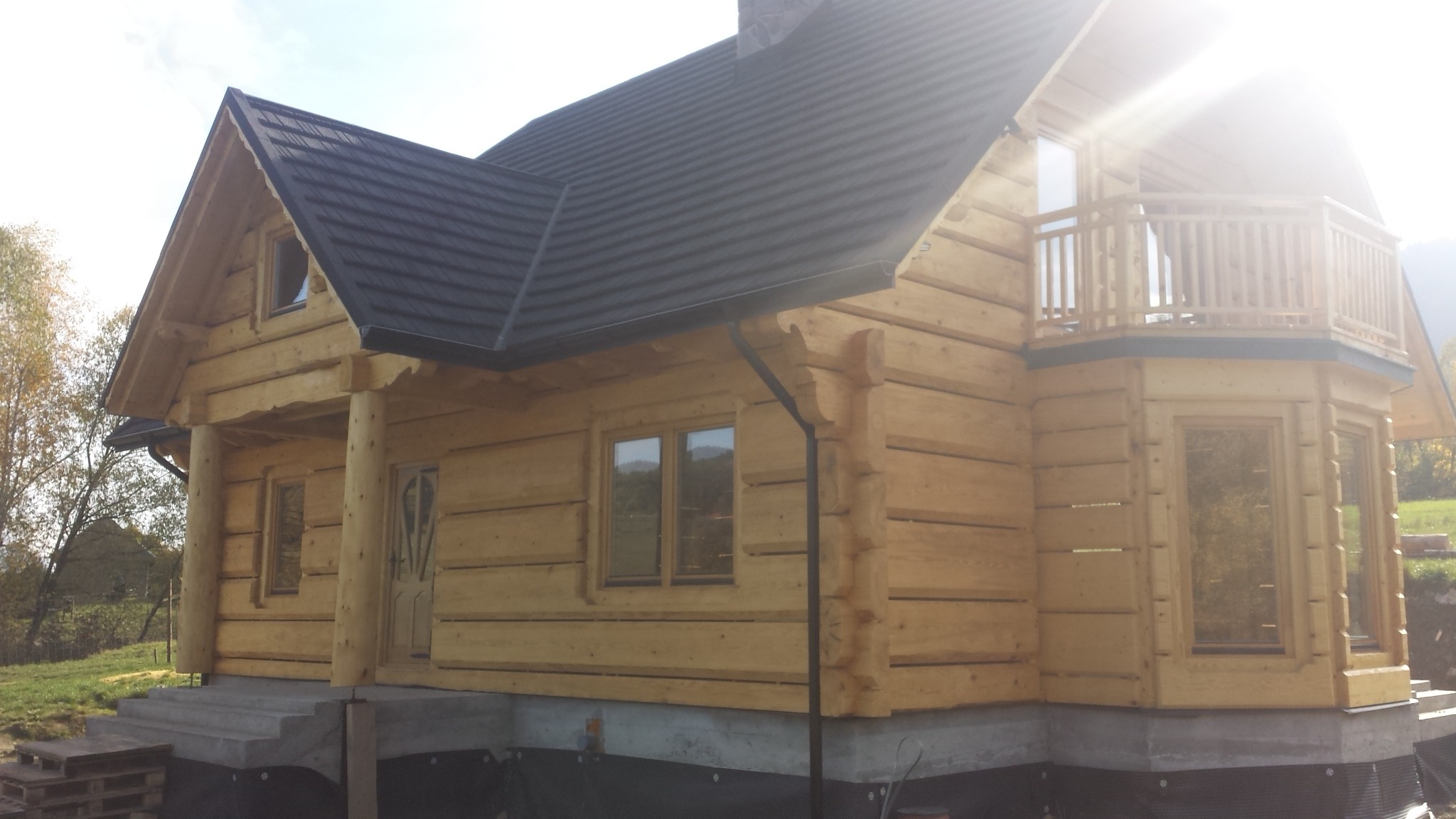 Nowoczesny dom z bali z balkonem, czarnym dachem i widoczną strukturą drewna, osadzony na fundamencie z izolacją, w słoneczny dzień.