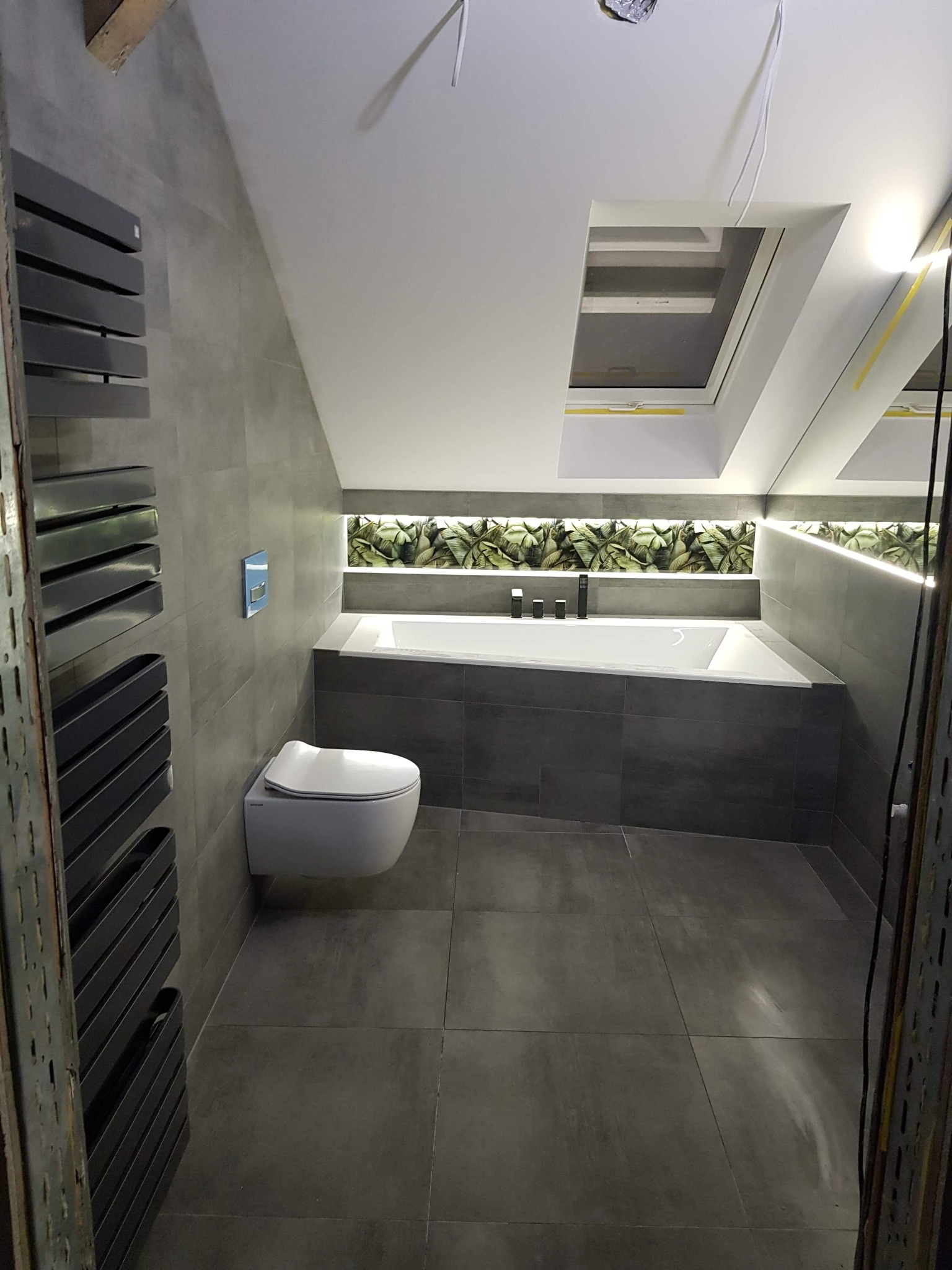 Nowoczesna łazienka na poddaszu z szarą glazurą, podświetlaną wanną, toaletą podwieszaną i grzejnikiem drabinkowym. Okno dachowe doświetla wnętrze.