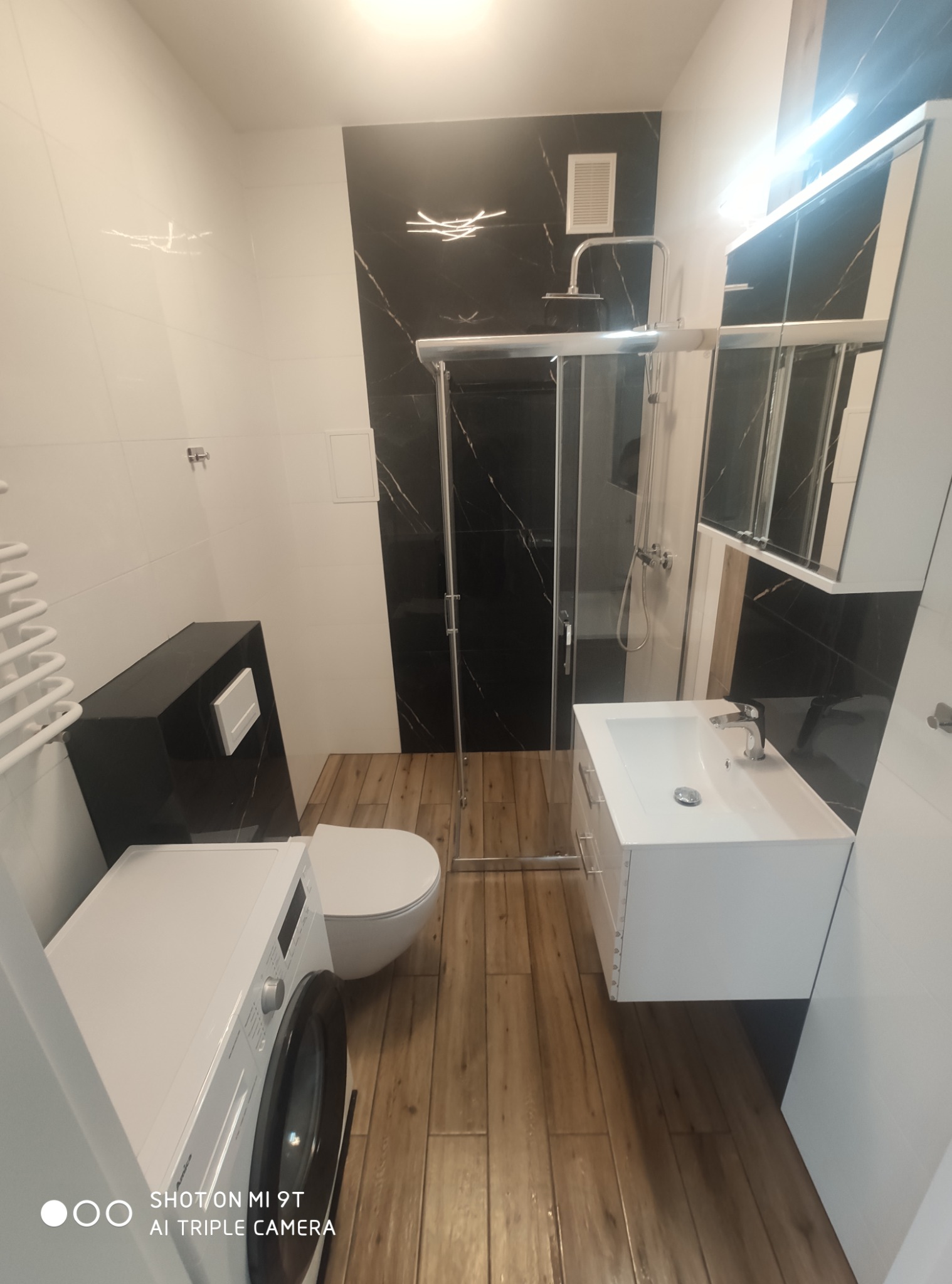 Nowoczesna łazienka po remoncie: kabina prysznicowa ze szklanymi drzwiami, umywalka z szafką, toaleta i pralka. Na ścianach białe i czarne płytki imitujące marmur, podłoga drewnopodobna.