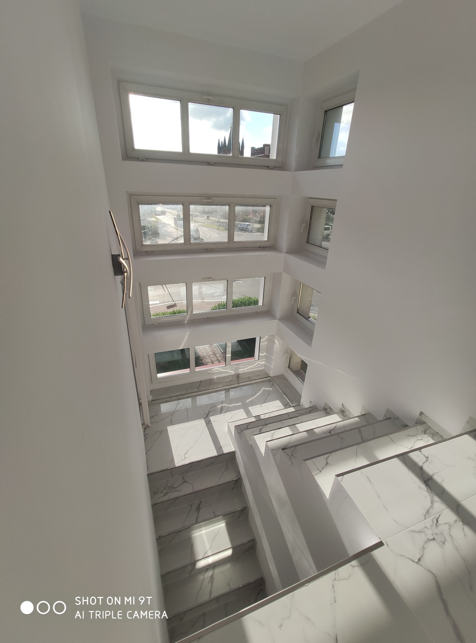 Białe schody z marmurowym wzorem w jasnym, nowoczesnym wnętrzu z oknami na kilku poziomach. Widok z góry.