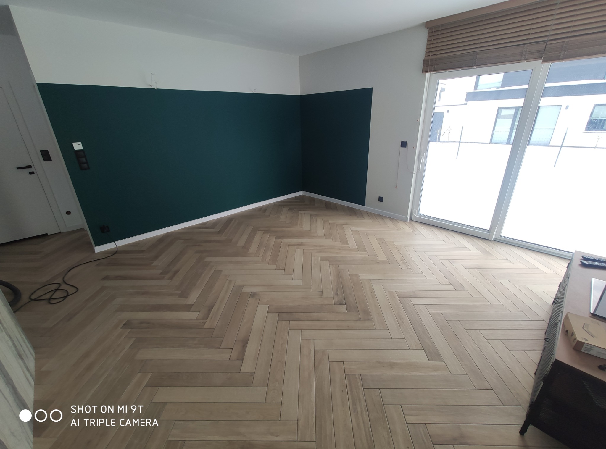 Pokój po remoncie: parkiet w jodełkę, ściana podzielona na białą górę i ciemnozielony dół. Duże okno z żaluzjami, minimalizm, nowoczesny design.