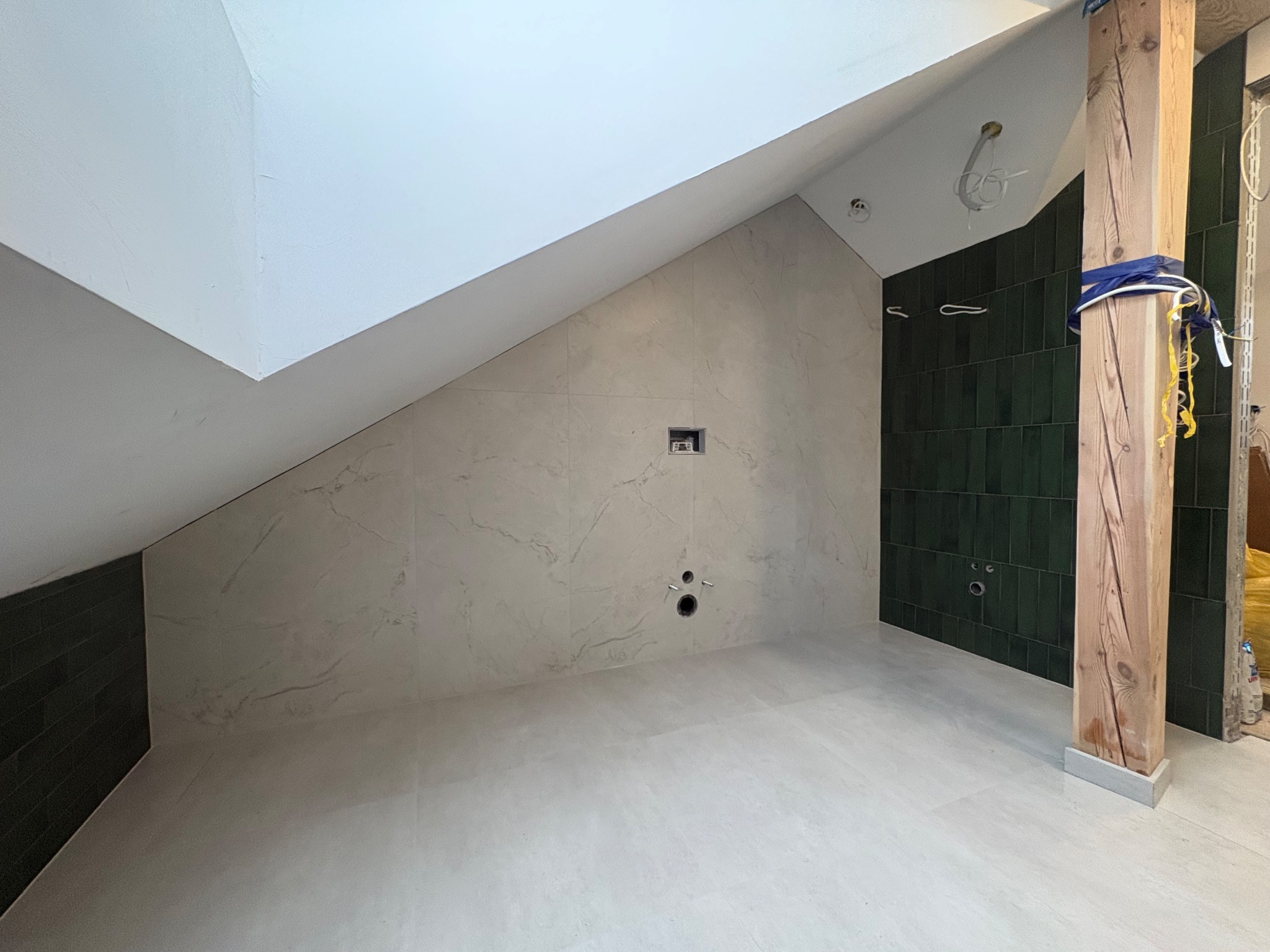 Wykończona łazienka na poddaszu z beżowymi i zielonymi płytkami, widoczne przyłącza hydrauliczne i drewniany słup. Ukończone podłogi i ściany.