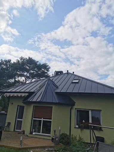 Dom z ciemnoszarym dachem, widoczne okno dachowe i nietypowa wieżyczka. Jasnozielona elewacja, błękitne niebo z chmurami w tle. Ujęcie z dołu, podkreślające geometrię dachu.