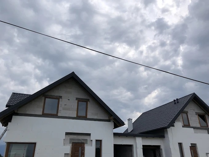 Dwa domy w trakcie budowy z szarymi dachami i białymi ścianami, widoczne okna i drzwi, pochmurne niebo w tle. Jeden dom z ociepleniem, drugi w stanie surowym.