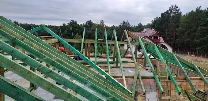 Wznoszenie drewnianej konstrukcji dachu z zielonych belek na tle lasu i domu z czerwoną dachówką. Widok z góry na budowę w trakcie realizacji.