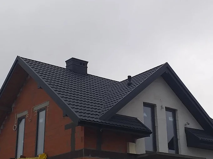 Dom w trakcie budowy z ciemnym dachem z dachówki, widoczne okna i komin. Część budynku w surowym stanie, druga otynkowana na biało. Pochmurne niebo w tle.