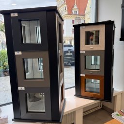 Globi Okna Małgorzata Juszczak - Ekspozycja okien z różnymi ramami i wykończeniami w salonie, na tle okna z widokiem na zabytkową architekturę miejską. Prezentacja wzornictwa i kolorystyki.