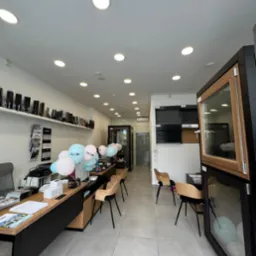 Wnętrze salonu z ekspozycją okien. Widoczne biurko z dekoracjami, krzesła oraz próbki okien z różnymi wykończeniami. Jasne oświetlenie i minimalistyczny design.