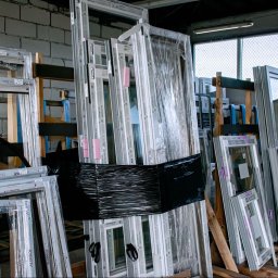 Globi Okna Małgorzata Juszczak - Magazyn okien i ram okiennych różnych producentów (Aluprof, Salamander) składowanych na drewnianych paletach. Część zafoliowana, widoczne etykiety i notatki.