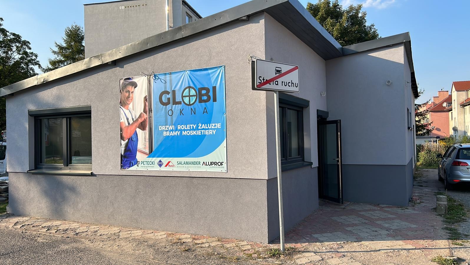 Szary budynek z reklamą firmy Globi Okna oferującej drzwi, rolety, żaluzje, bramy i moskitiery. Widoczne okno z roletą zewnętrzną i znak drogowy 'strefa ruchu'.
