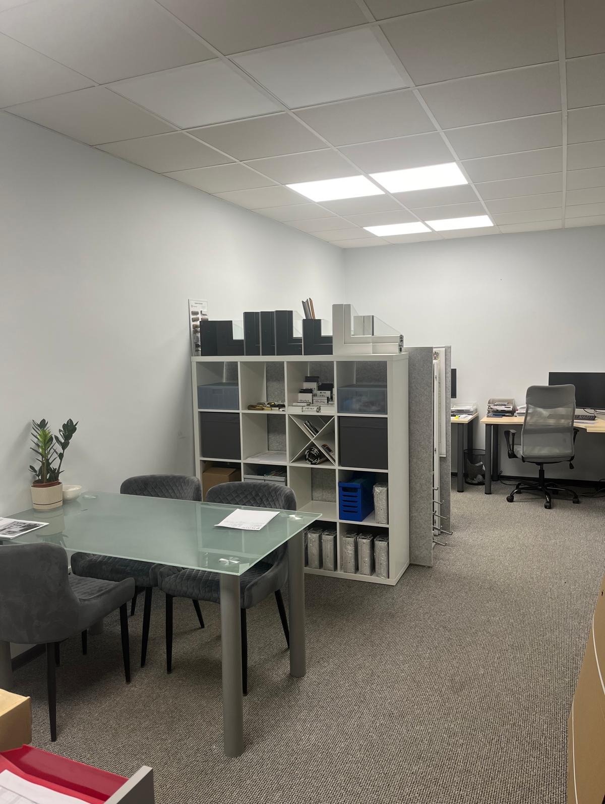 Jasne biuro z przeszklonym stołem i regałem z próbkami profili okiennych. Widoczne biurka w tle. Przykładowe profile okienne na regale.