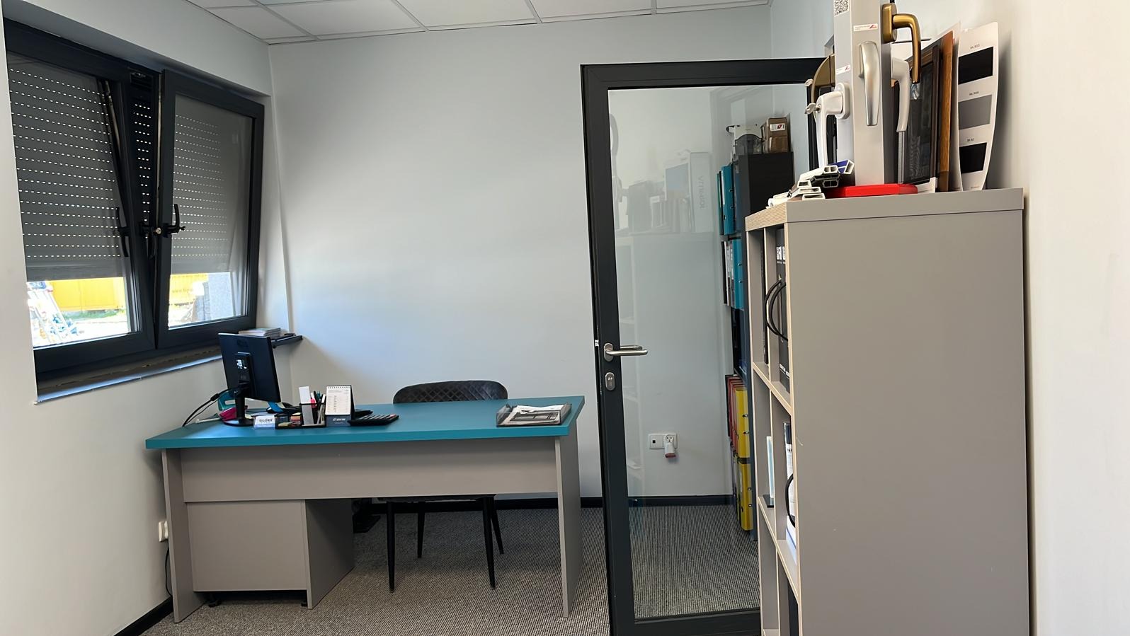 Biuro z oknem z roletami zewnętrznymi, drzwiami z czarną ramą i regałem z próbkami klamek i kolorów. W tle widoczny fragment żółtej elewacji budynku.