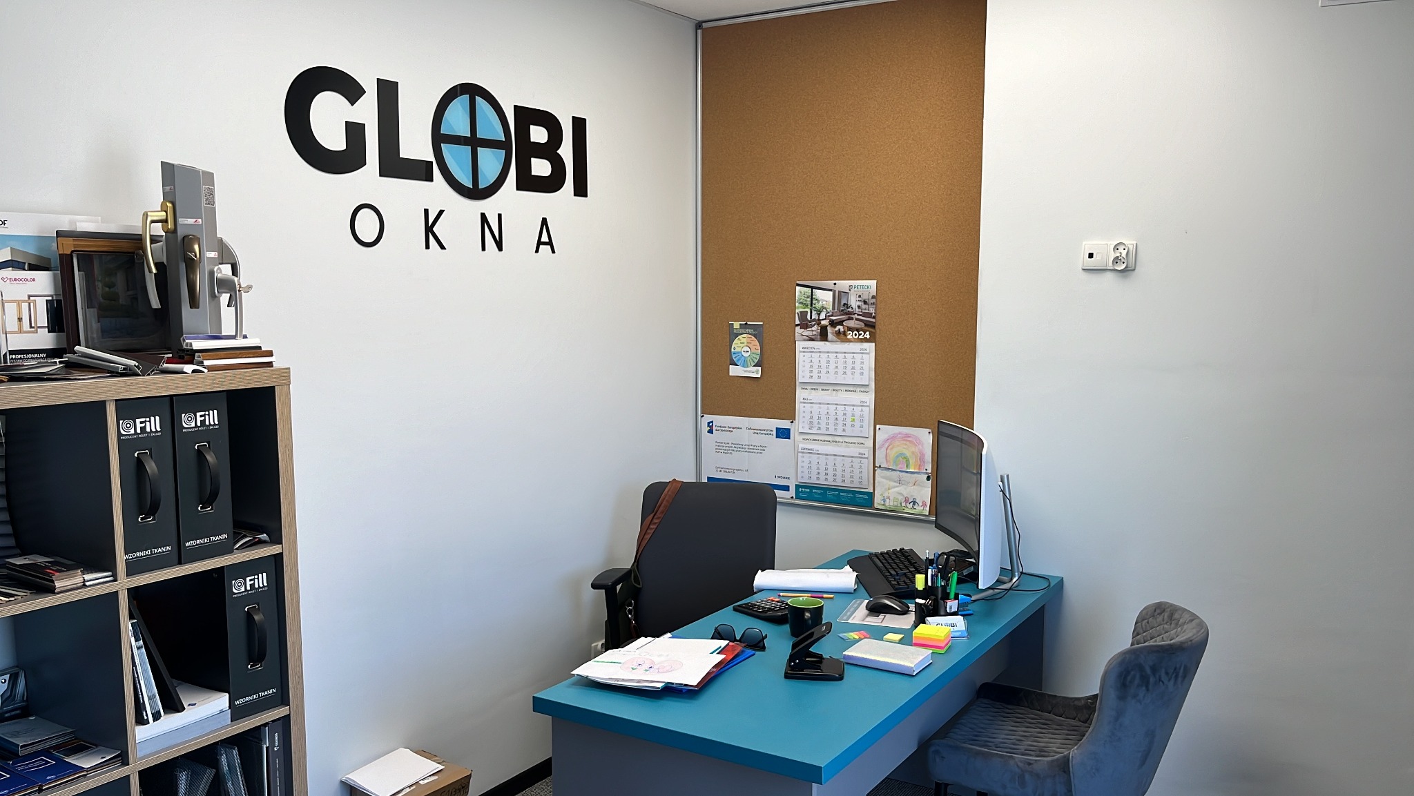 Biuro firmy z logo 'Globi Okna', ekspozycją próbek i katalogów. Widok na biurko z komputerem, kalendarzem i materiałami biurowymi. Jasne wnętrze, minimalistyczny design.