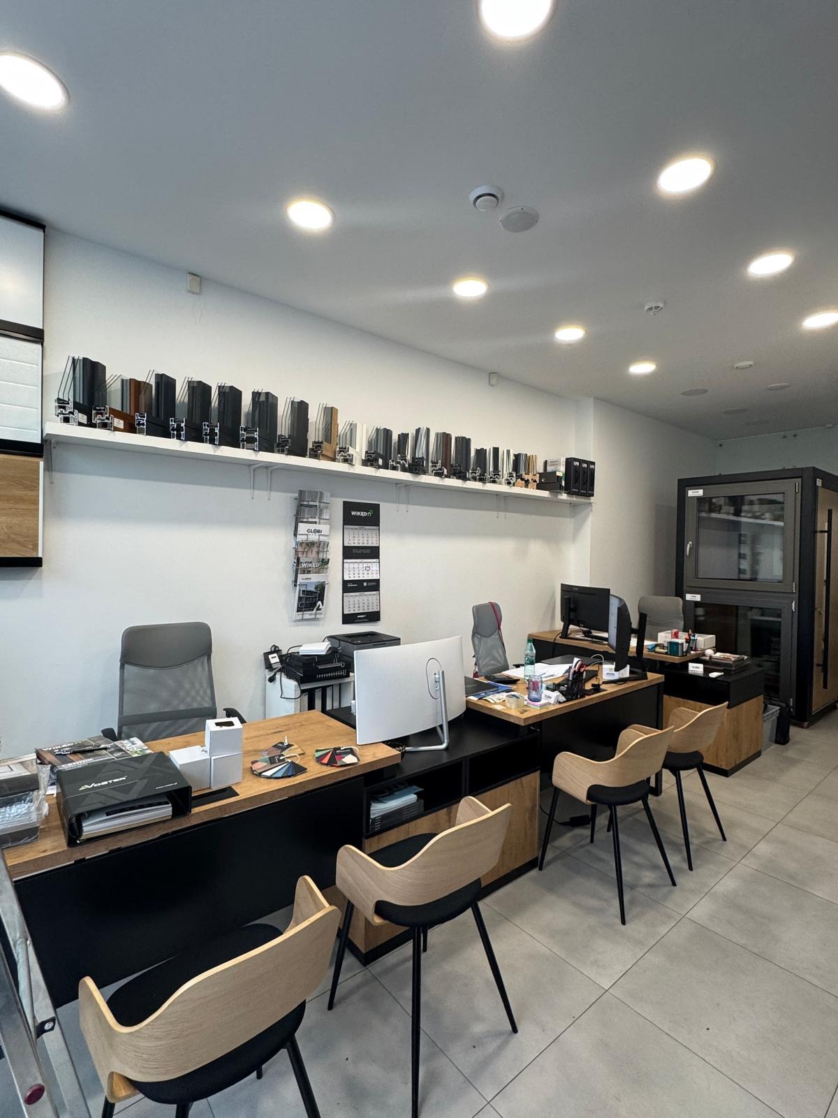 Wnętrze biura z ekspozycją profili okiennych na ścianie. Na pierwszym planie biurka, krzesła i katalogi. W tle widoczny model okna.